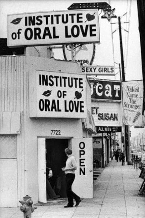 A person standing outside the Institute of Oral Love with signs promoting it, along a sidewalk with other storefronts and signs in the background, including one for 'Sexy Girls' and 'Naked Came The Stranger'.