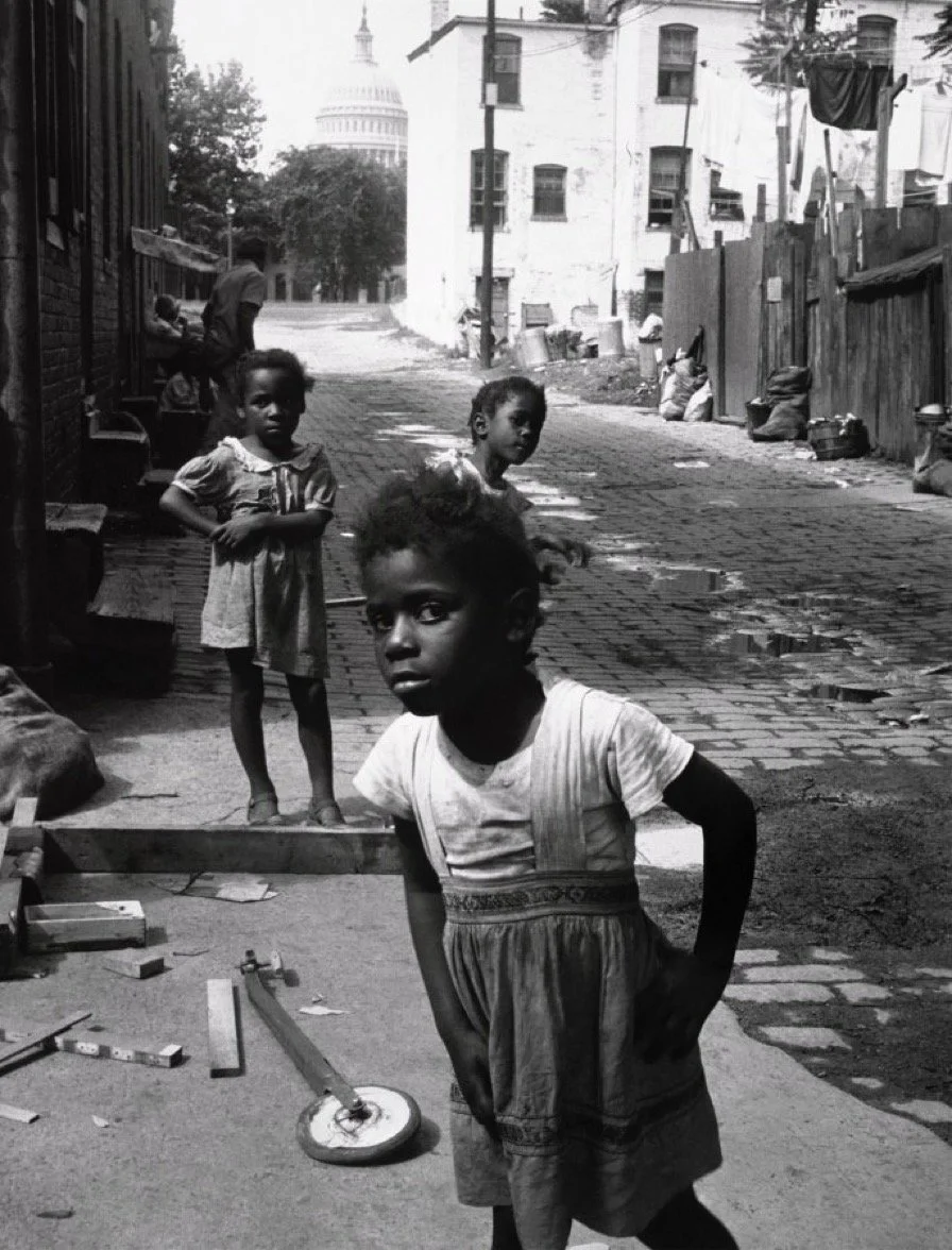 Four children standing on a dirt and cobblestone street in an impoverished neighborhood, with the United States Capitol visible in the distance.