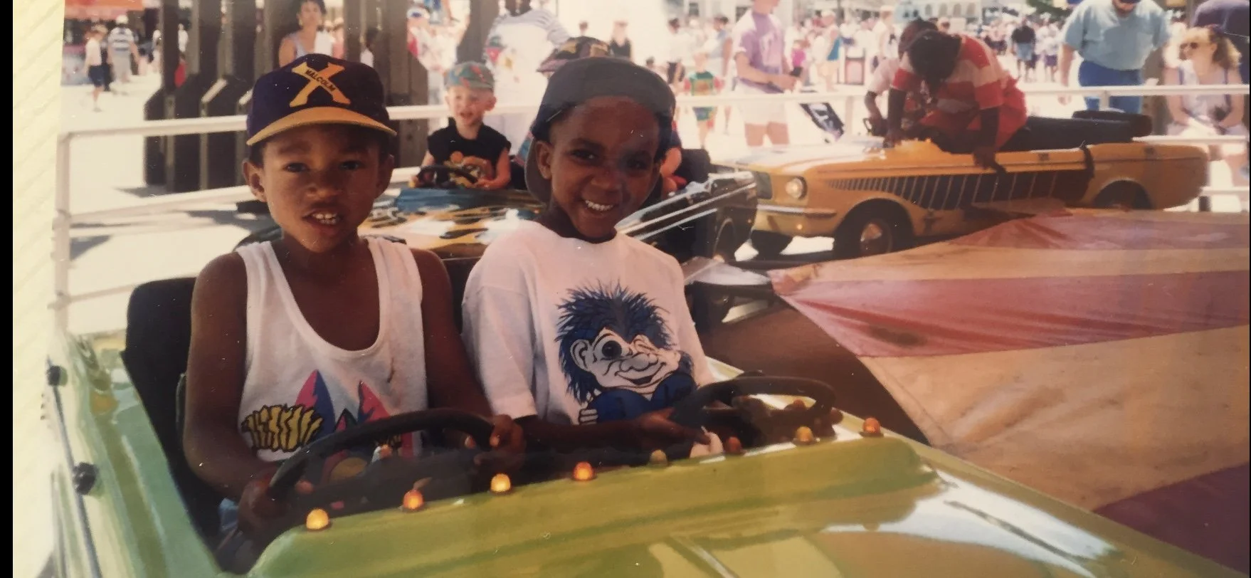 Two boys are excited in amusement park rides, sitting in a small colorful car ride, with other children and classic cars in the background.