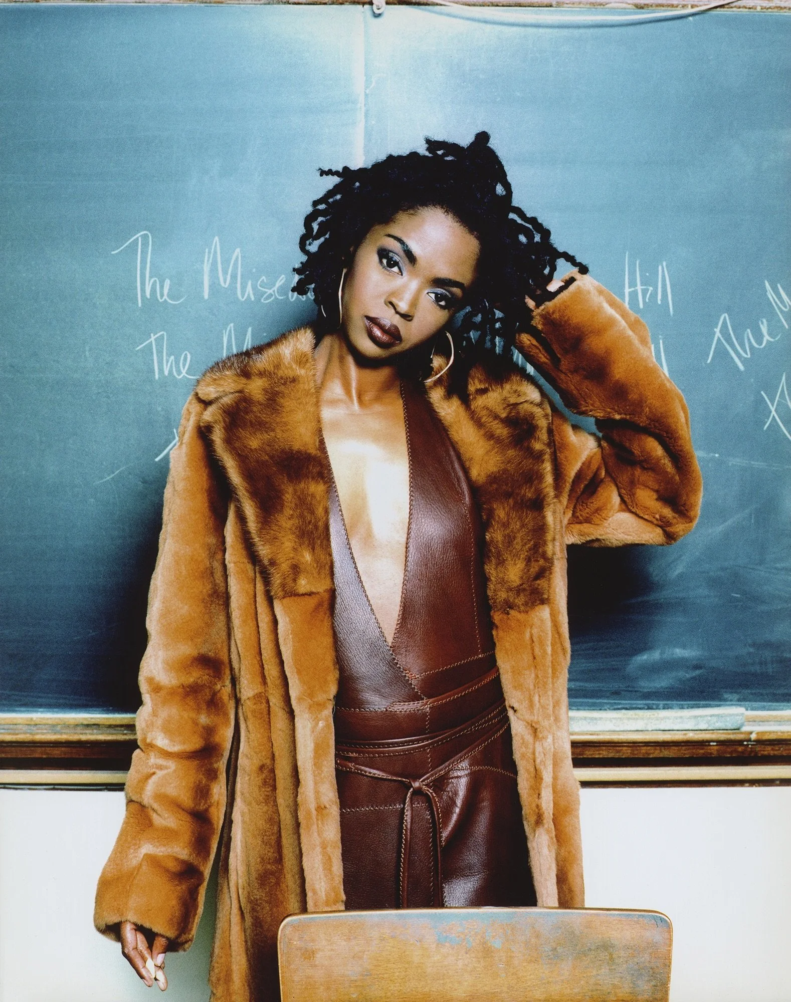 A woman with dark beauty and curly hair standing in front of a blackboard, wearing a brown leather dress and a tan fur coat.
