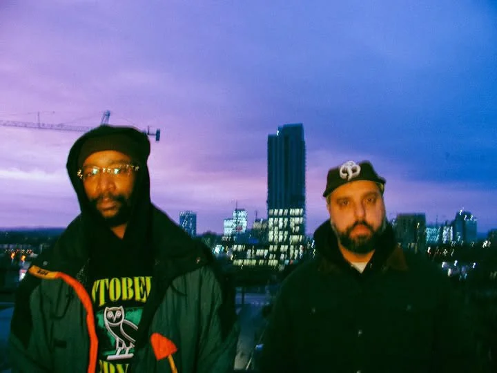 Two men standing on a rooftop at dusk with city skyline and tall buildings in the background.