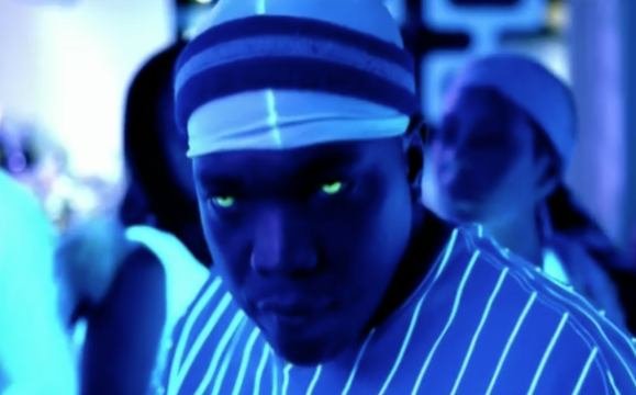 A person with a striped head covering and striped shirt, looking intensely at the camera, in a blue-lit environment with other people in the background.