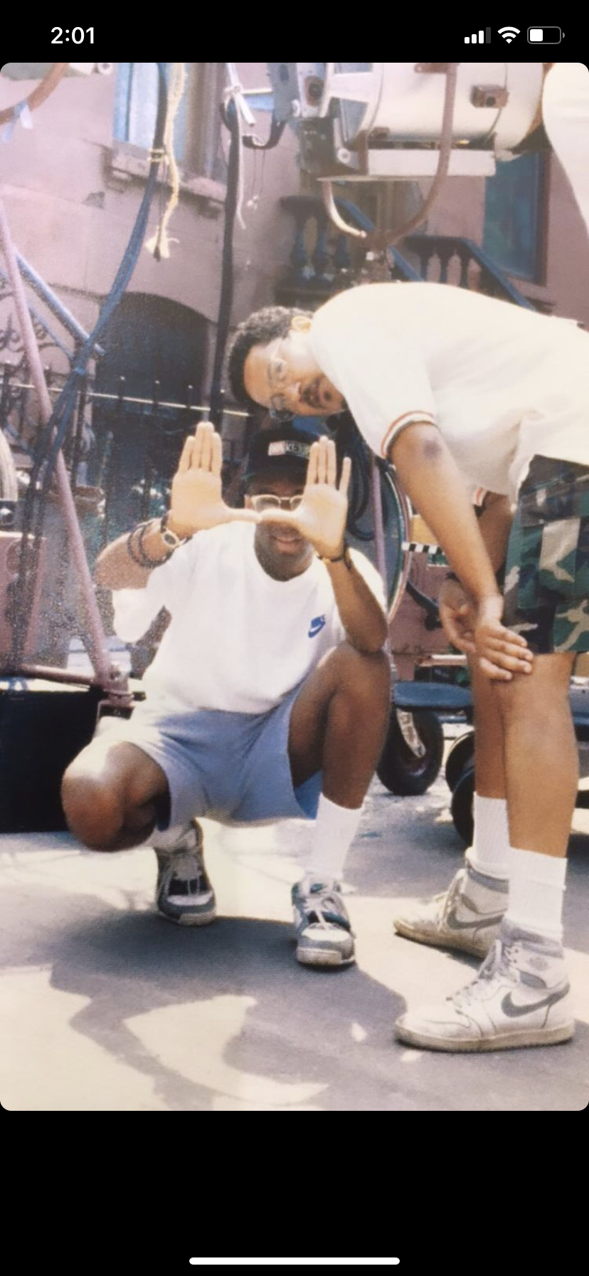 Two young men posing outdoors; one is crouching with hands framing his face, smiling, wearing a white t-shirt, gray shorts, and a red cap, the other is standing, leaning over, with glasses and face paint, wearing a white polo shirt and camouflage sho