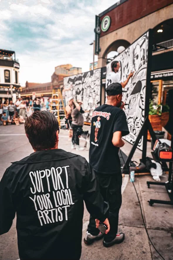 People painting murals outdoors at a street event with onlookers, buildings, and a parking sign in the background.