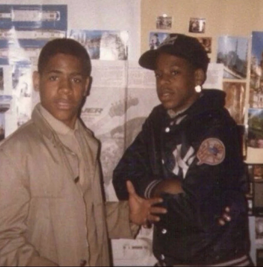 Two young men standing indoors, with one shaking hands with the other. The background shows various posters and images on the wall.