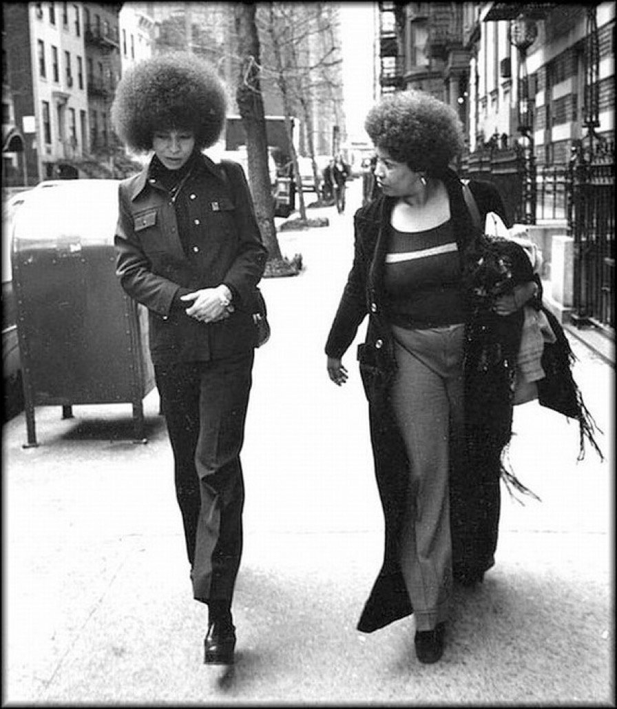 Two women with large afro hairstyles walking down a city sidewalk, dressed in vintage fashion, with buildings and pedestrians in the background.