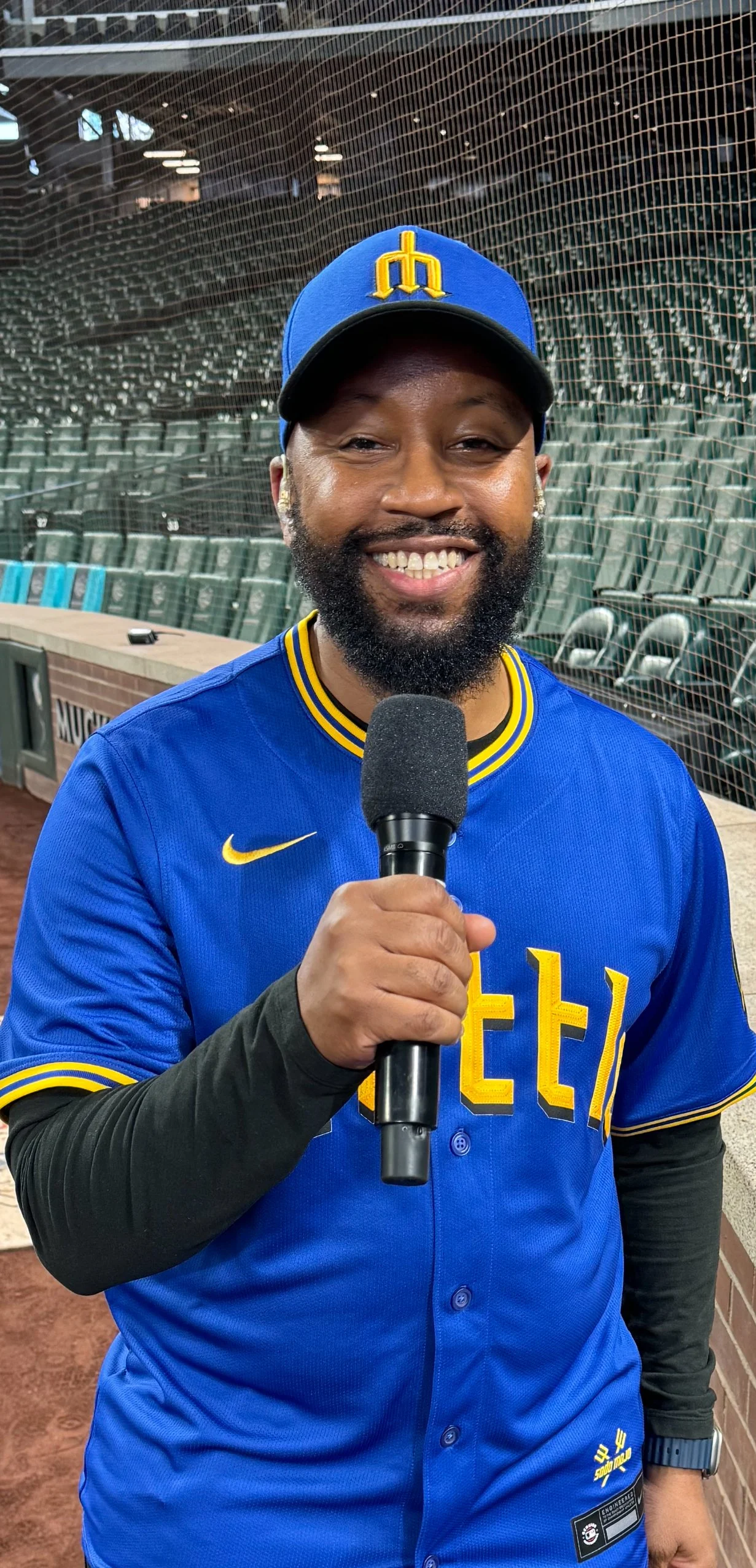 Person wearing a blue and yellow baseball jersey with a matching cap, holding a microphone, standing in a baseball stadium with empty seats.