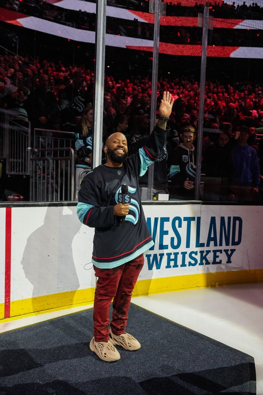 A man wearing a Seattle Kraken hockey jersey waving and holding a microphone on an ice rink platform. The background shows a crowd and the partially visible "WESTLAND WHISKEY" advertisement.