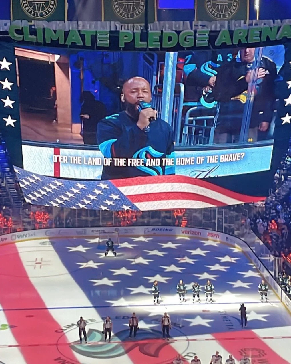 Singer performs national anthem at Climate Pledge Arena before a hockey game, with American flag displayed on screen and ice.