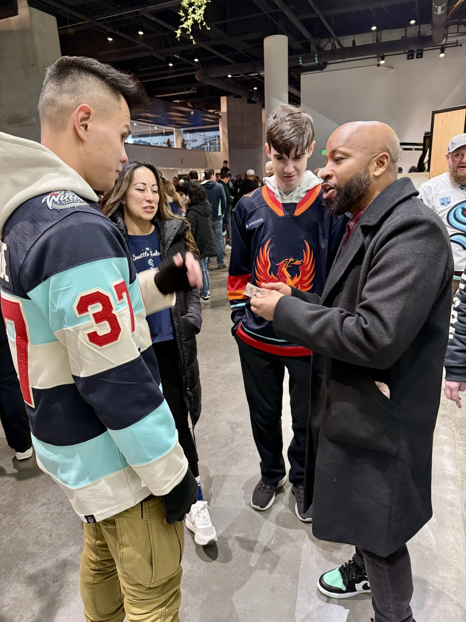 A group of people having a conversation indoors. One person is wearing a light blue hockey jersey, another a dark blue hockey jersey. They are all casually dressed and standing in what appears to be a public space, possibly at an event.