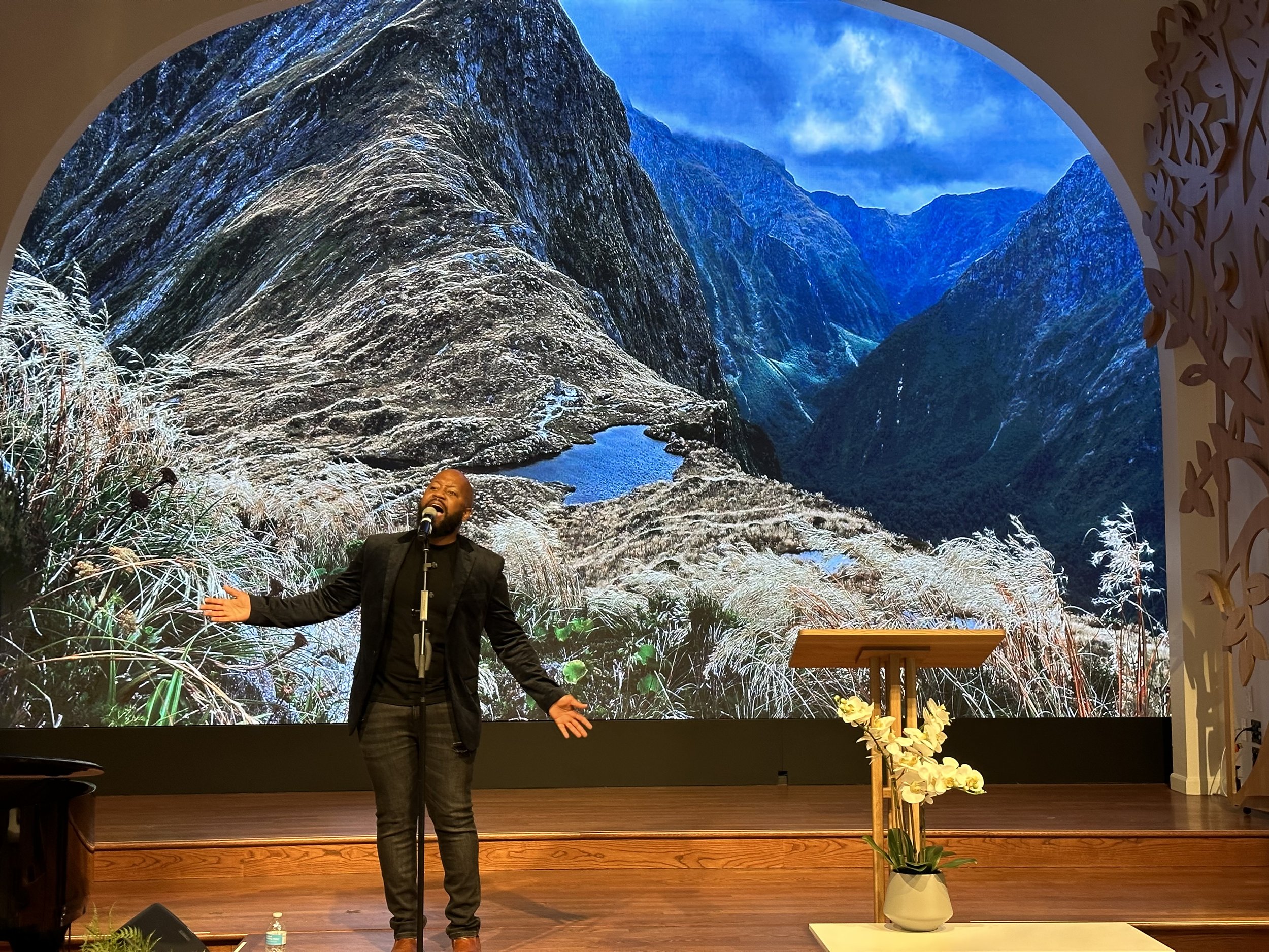 A person passionately singing with arms open on a stage in front of a large screen displaying a mountain landscape with a valley and a small lake. The stage has wooden features and a lectern with white flowers in a vase.