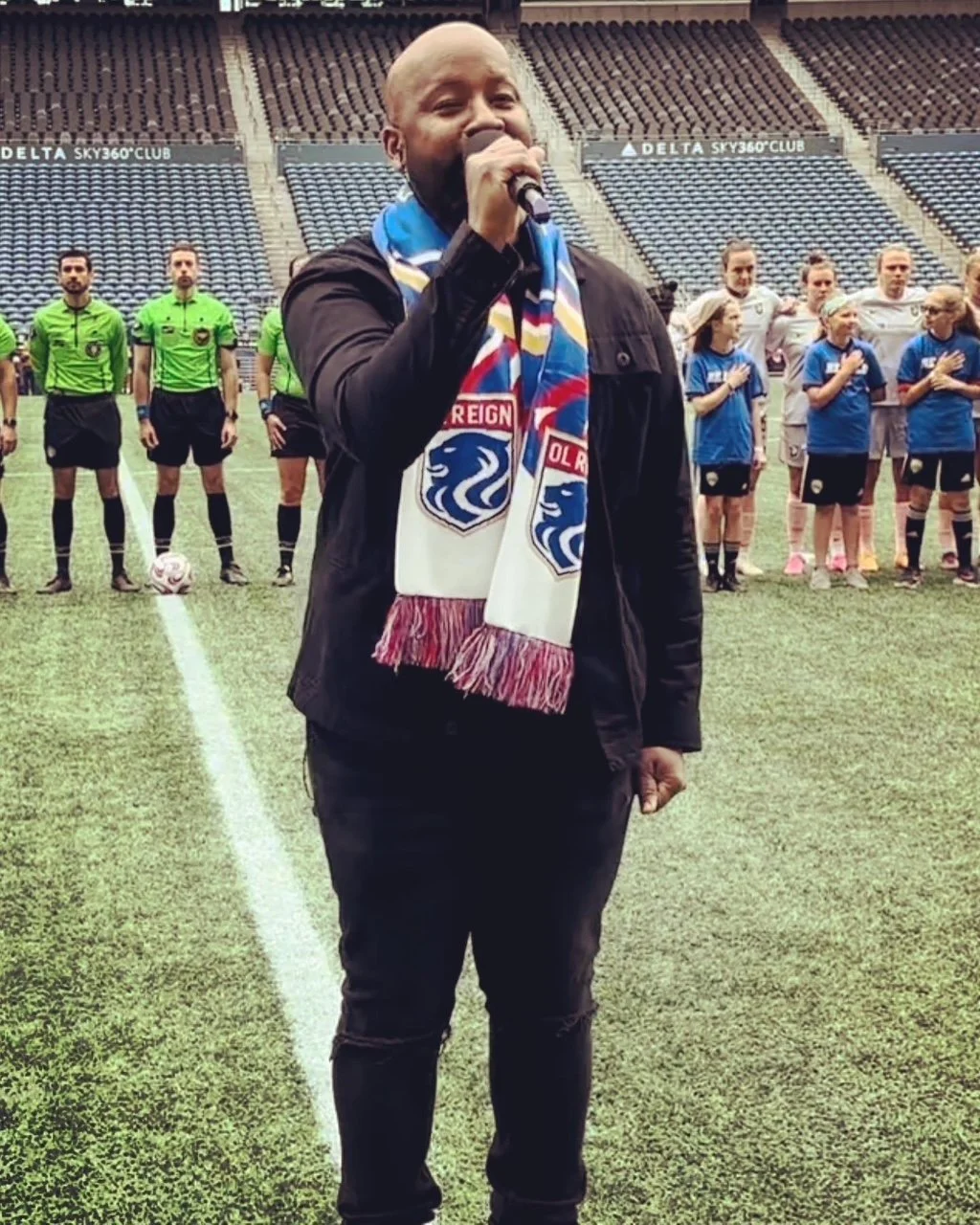 A person wearing a colorful scarf holds a microphone and sings at a sports event, with sports officials and players standing in the background on the field.