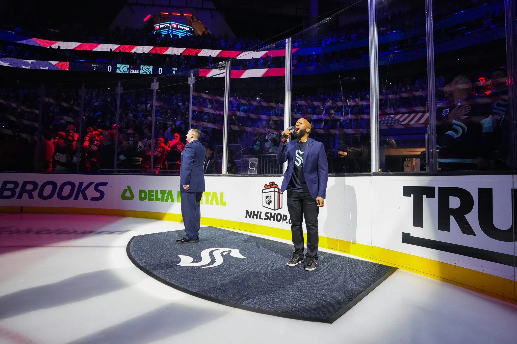 A man in a blue jacket sings into a microphone on an ice hockey rink next to a man in a suit. The American flag is displayed above, and players with hockey uniforms are visible in the background. The rink is lined with advertisements and the logo of 