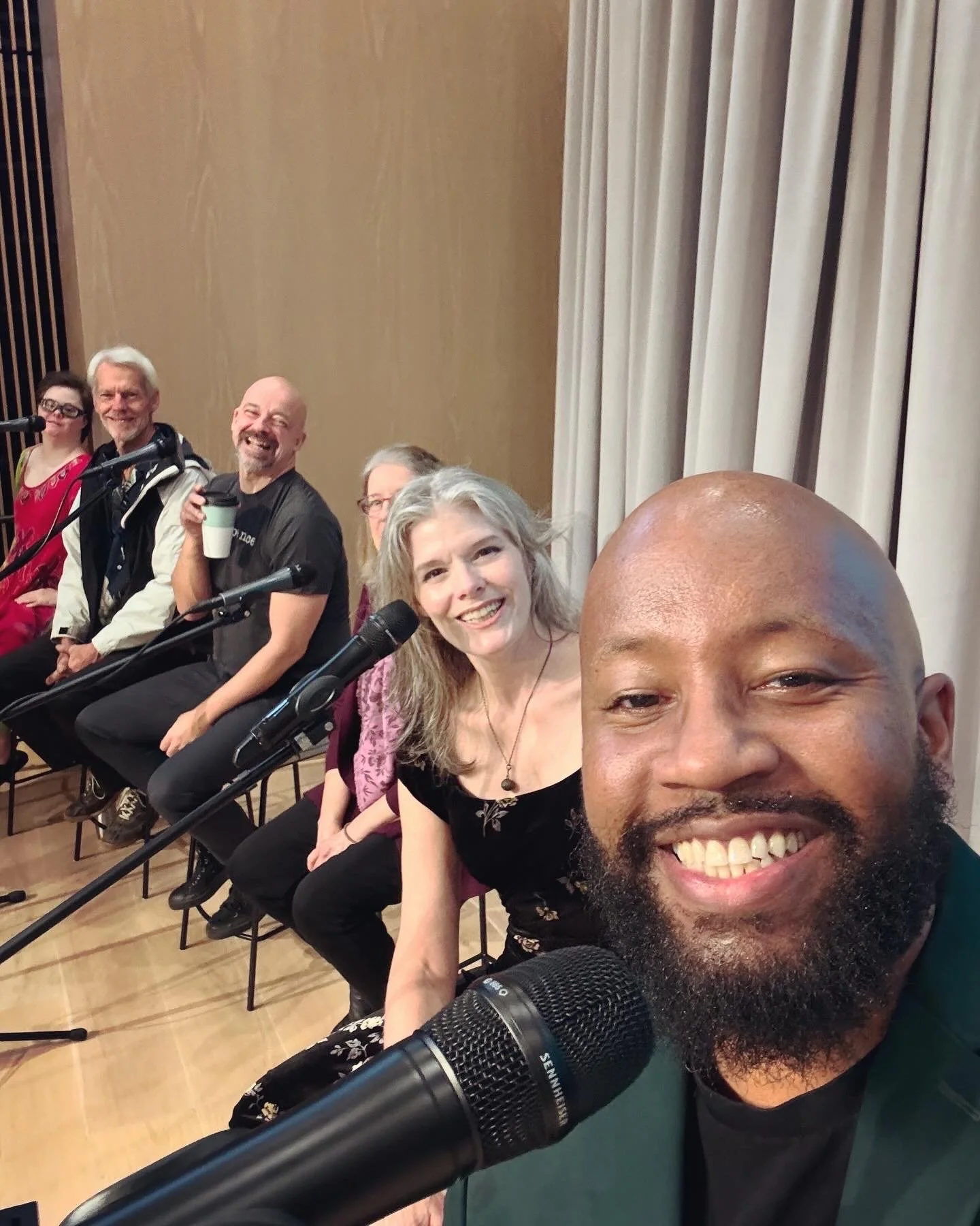 Group of six people sitting on stage with microphones, one taking a selfie