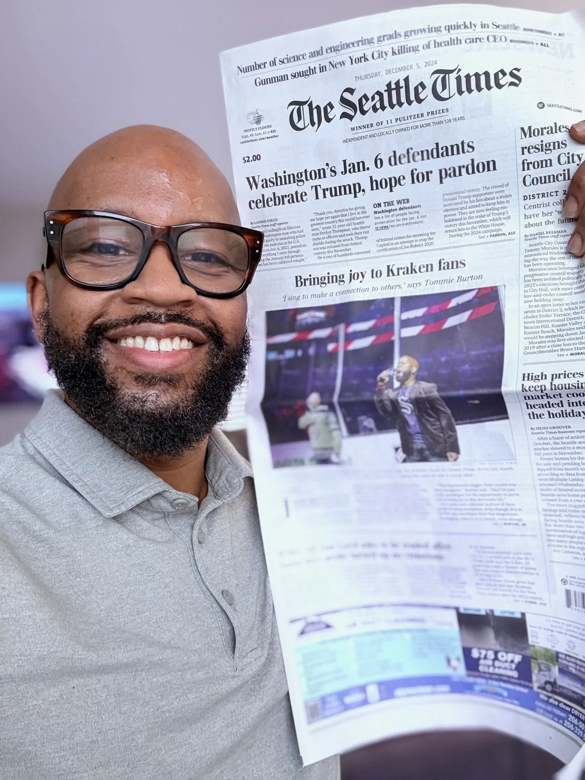 A person with glasses smiling and holding a newspaper titled "The Seattle Times" with a headline about Jan. 6 defendants and Trump on the front page. The date on the newspaper is Thursday, December 5, 2024.