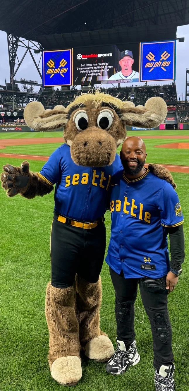 A person posing with a baseball team mascot on a field, wearing matching team jerseys in a stadium. The scoreboard displays a player’s image and stats in the background.