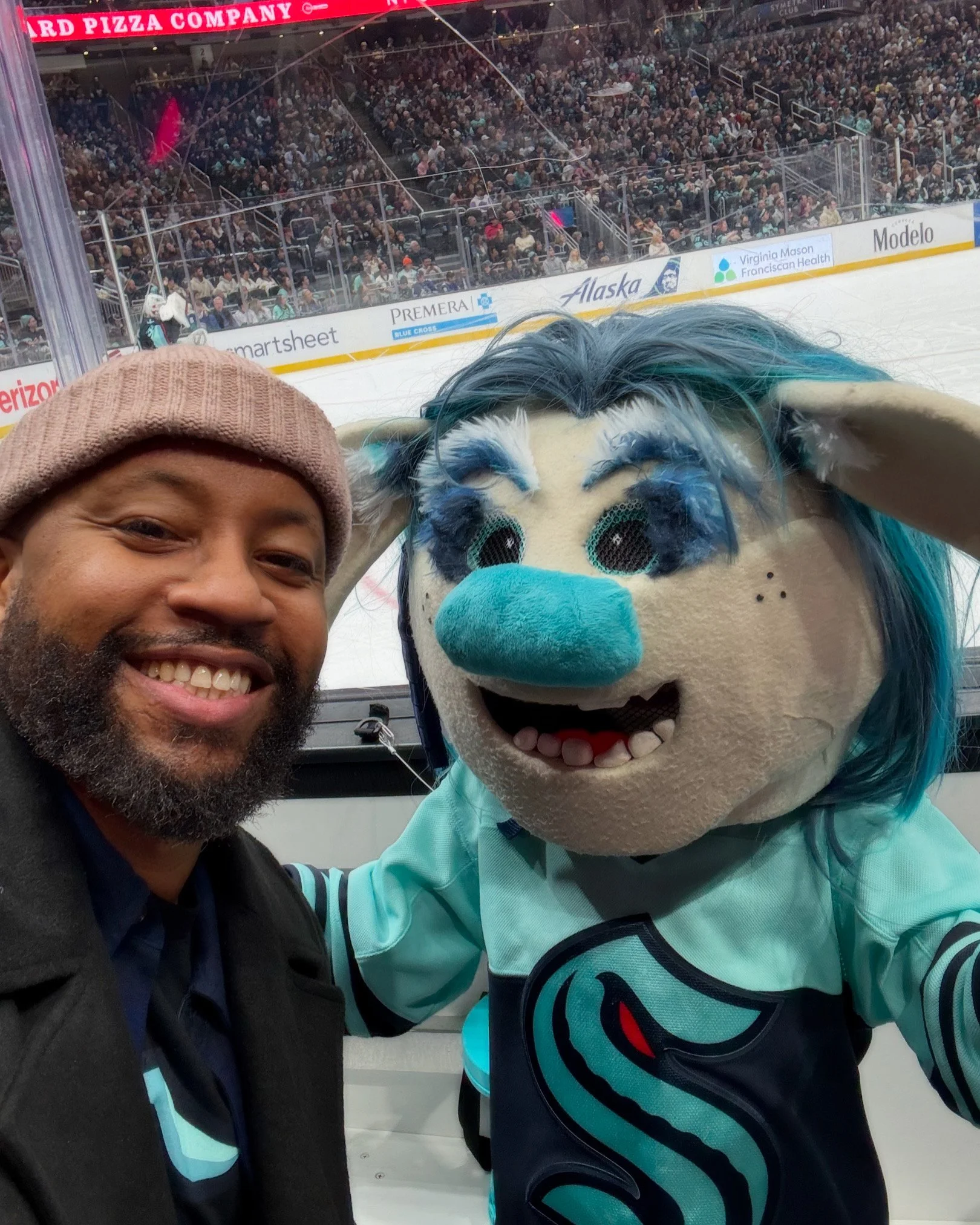 Person taking a selfie with a hockey team mascot at an ice rink.