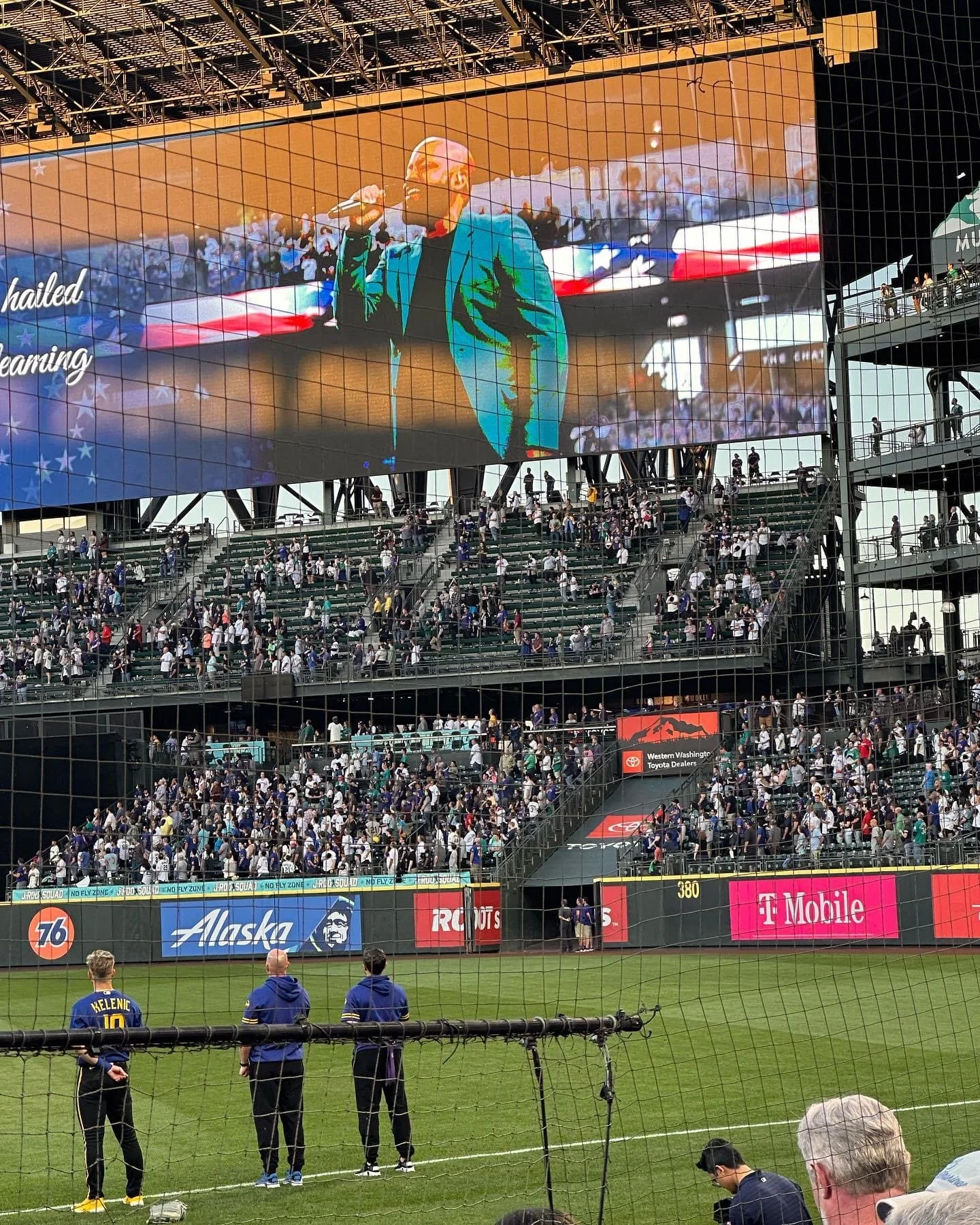 A large stadium screen displays a person singing the national anthem. The crowd stands in the background. Sports advertisements are visible along the stadium wall.