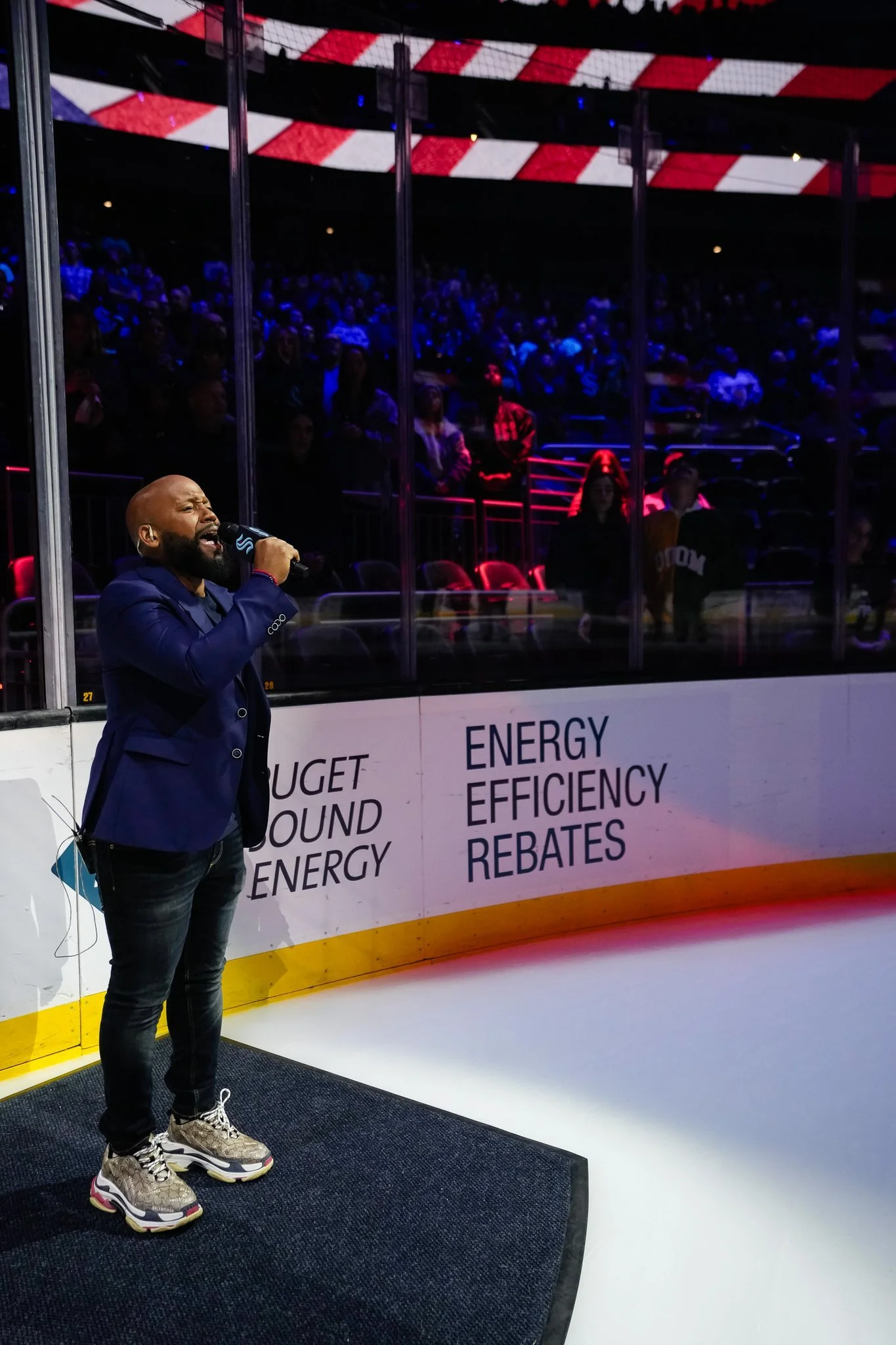Man in a blue blazer singing into a microphone on an ice rink beside a board with text 'ENERGY EFFICIENCY REBATES' and 'PUGET SOUND ENERGY,' in a stadium with audience visible in the background.