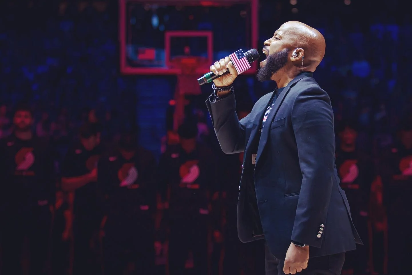 A man singing into a microphone covered with an American flag print at a basketball game, with players standing in the background.