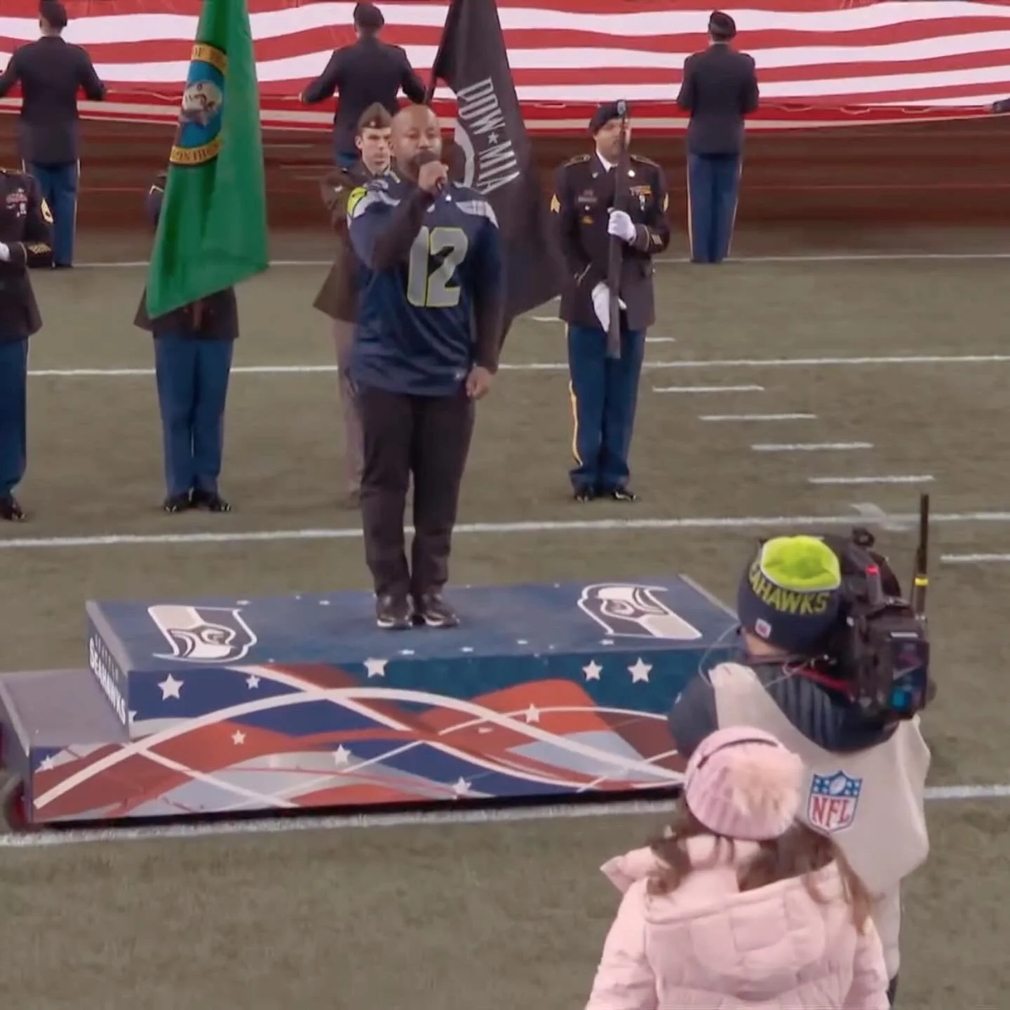 Man wearing a Seattle Seahawks jersey sings into a microphone on a decorated platform during a pre-game ceremony at a football field. Military personnel hold flags including the Washington state flag and a POW/MIA flag. A camera operator wearing an N