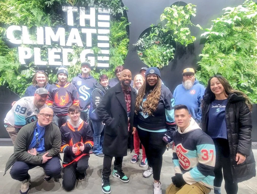 A group of people posing in front of a wall with "The Climate Pledge" written on it, surrounded by greenery. They are wearing hockey jerseys and smiling at the camera.