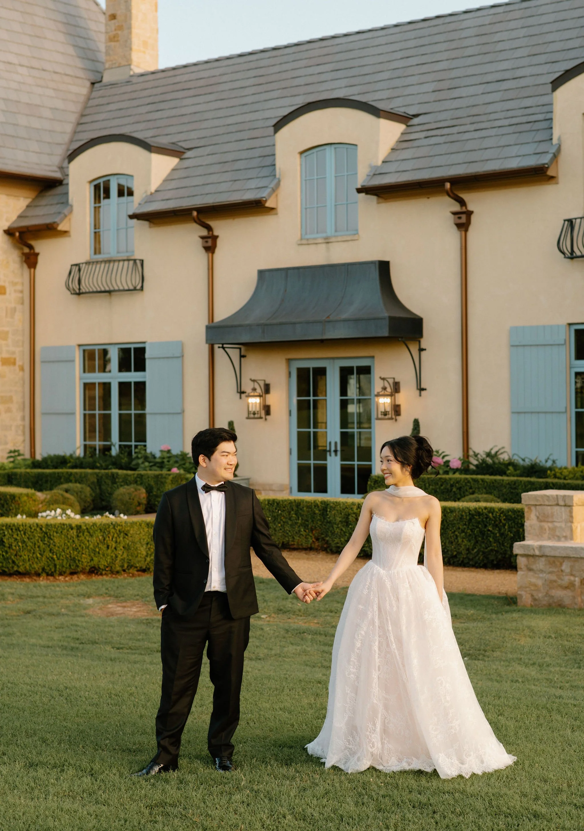 A bride and groom holding hands and smiling, standing on a lawn in front of a large house with blue doors and windows, and a slate roof.