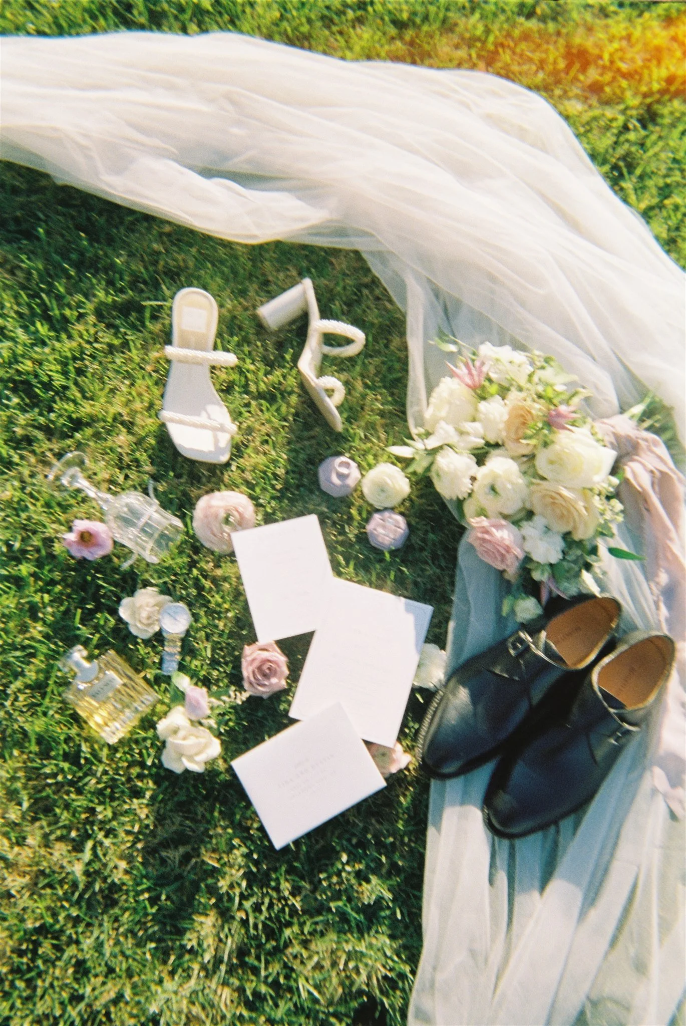A collection of white high-heeled shoes, a bouquet of white and pink flowers, perfume bottles, candles, handwritten notes, and a sheer fabric are arranged on the grass for a picnic-style wedding or romantic occasion.