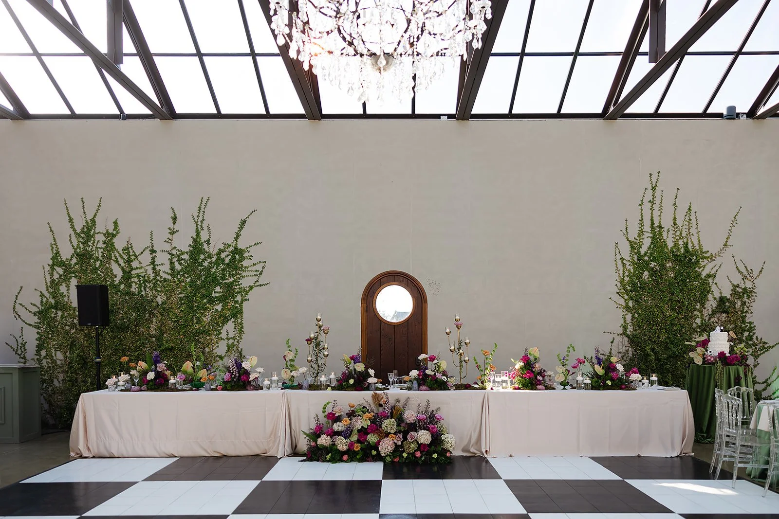 Wedding reception table with floral arrangements, candles, and a wooden door mirror in a modern indoor setting with a checkered black and white floor and a large chandelier overhead.