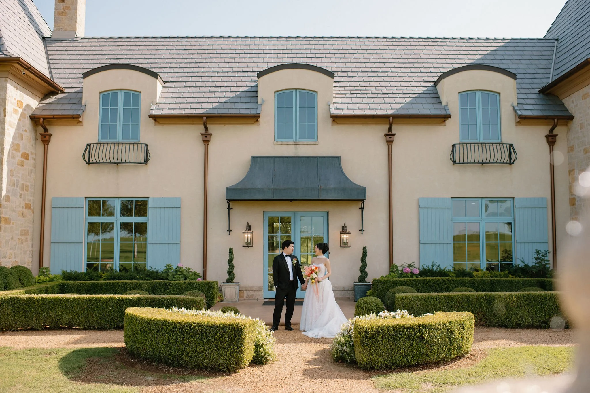 A bride and groom standing in front of a large house, holding hands and looking at each other. The bride is wearing a white wedding gown and holding a bouquet of flowers, while the groom is dressed in a black tuxedo. The house has beige walls, blue s