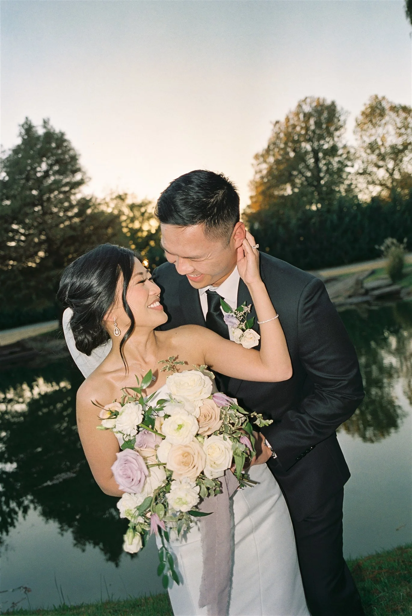 A newlywed couple smiling and embracing near a pond during sunset, with trees in the background.