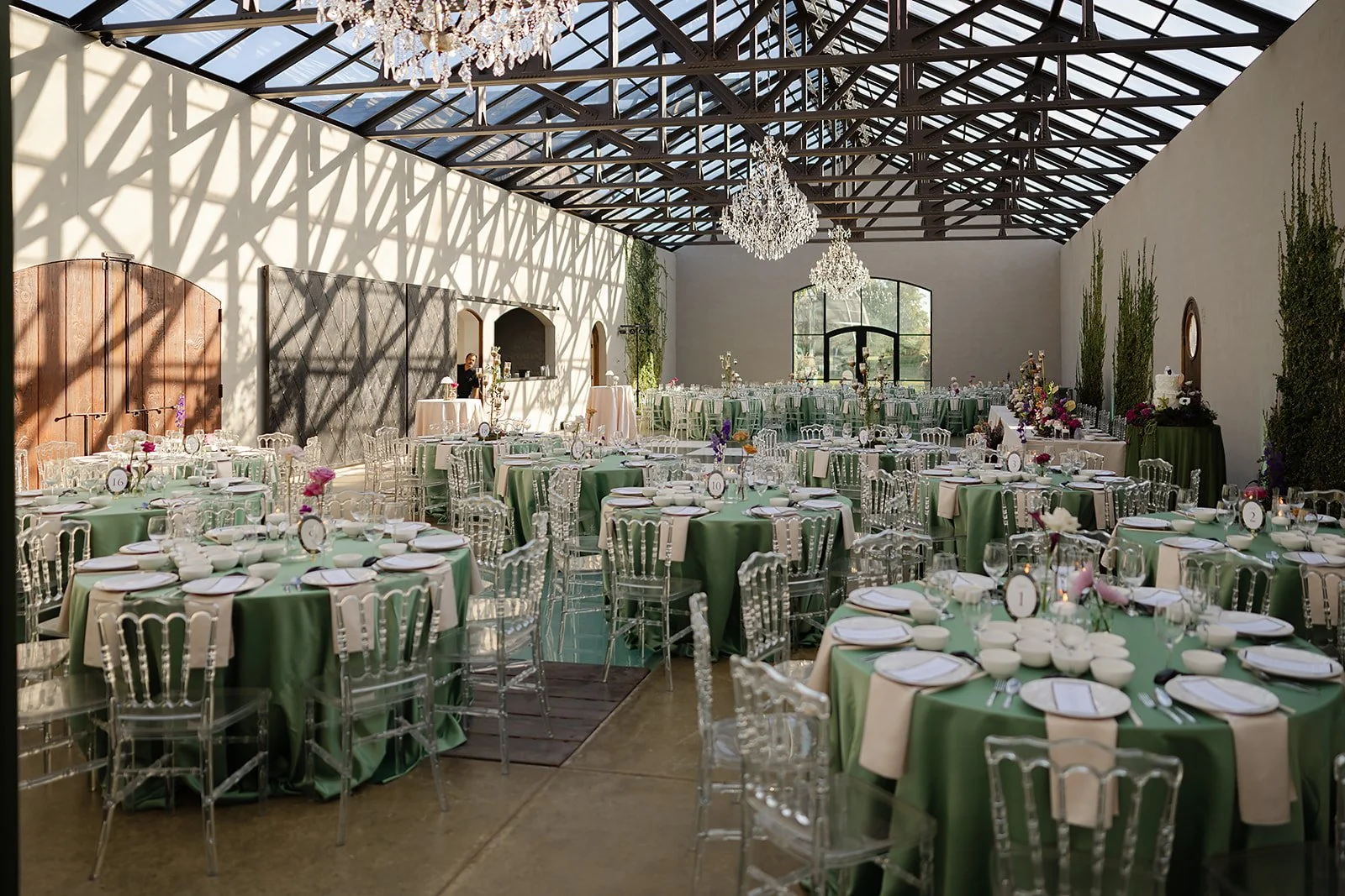 Elegant banquet hall with decorated tables covered in green and cream tablecloths, transparent chairs, floral centerpieces, and chandeliers hanging from a glass ceiling.