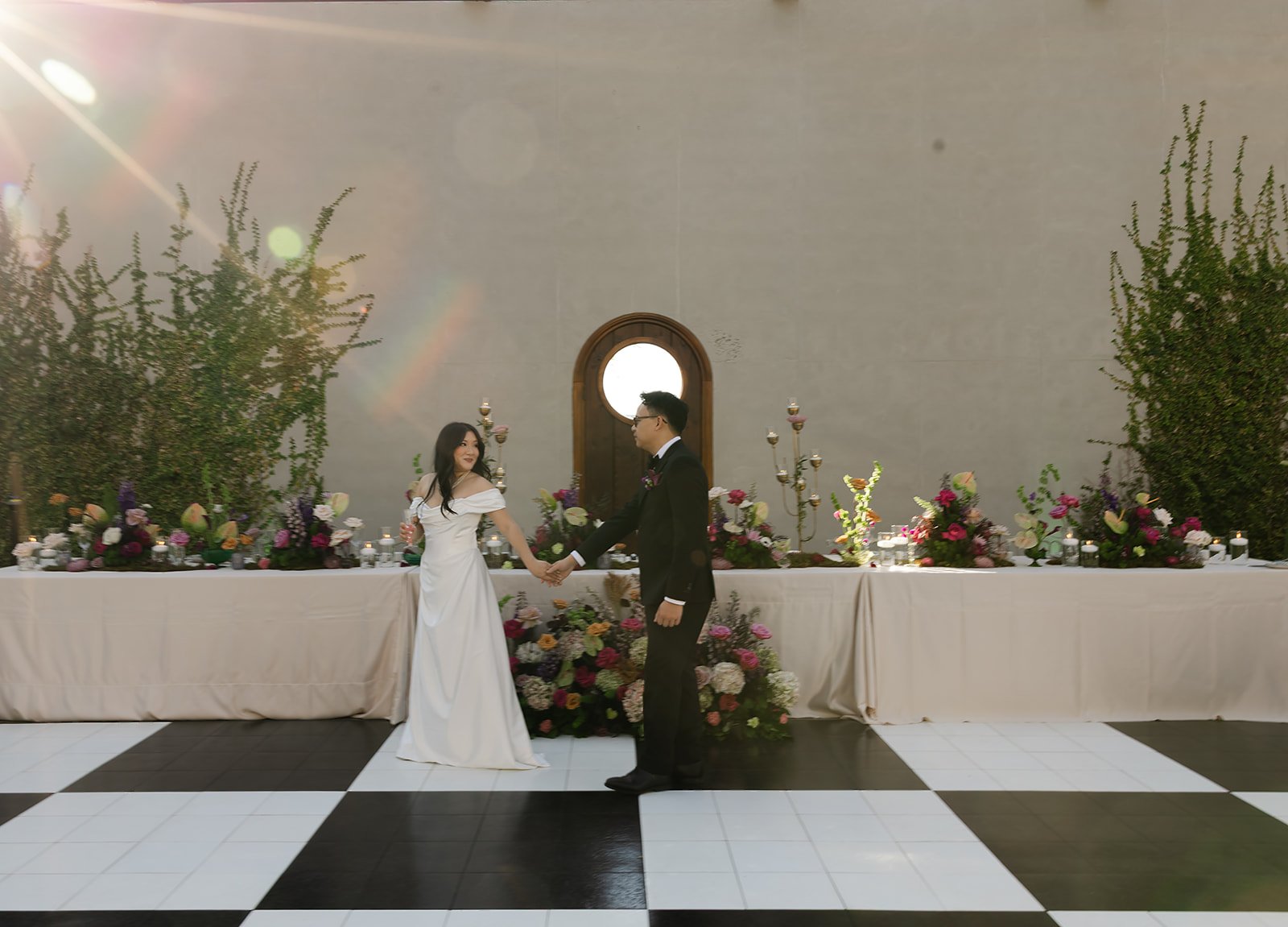 A bride and groom holding hands and smiling during their wedding ceremony in a decorated hall with flowers and candle arrangements, a large round window, and trees on either side.