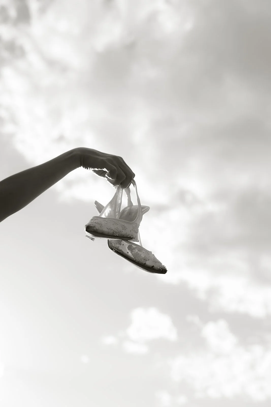 A hand holding a pair of wet flip-flops against a cloudy sky.