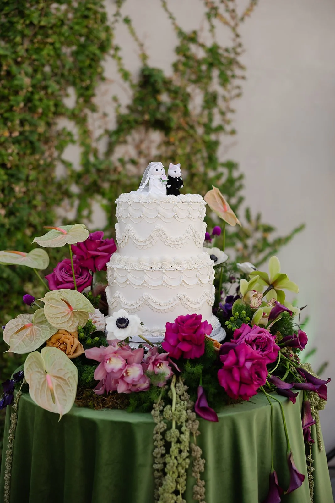 A two-tiered white wedding cake decorated with white icing swags and flowers, topped with bride and groom figurines of animals, surrounded by vibrant pink, purple, and cream flowers on a green tablecloth.