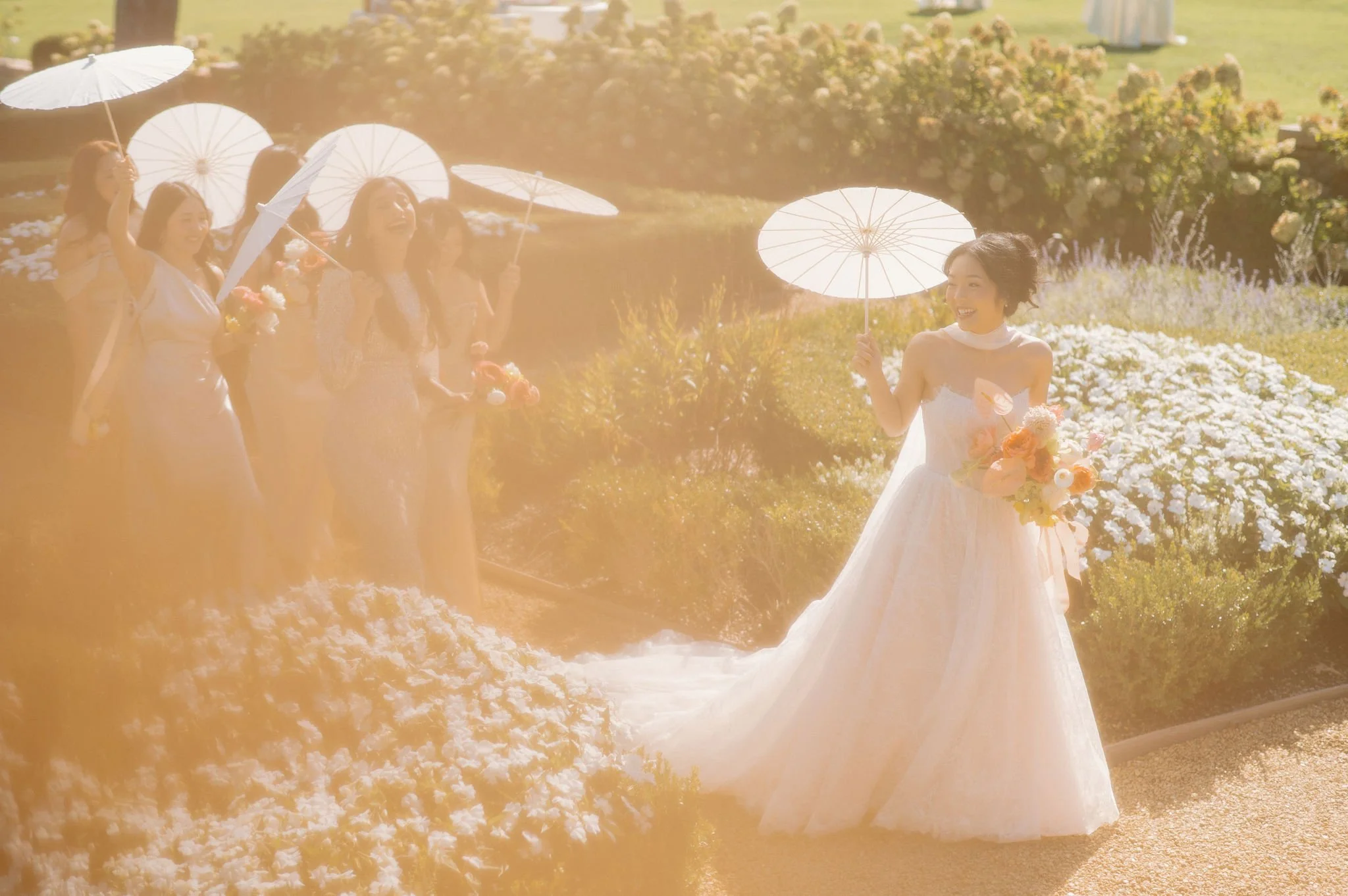 A bride in a white wedding dress holding flowers and a parasol, standing in a garden, smiling as a group of women in dresses holding parasols walk toward her, also in the garden on a sunny day.