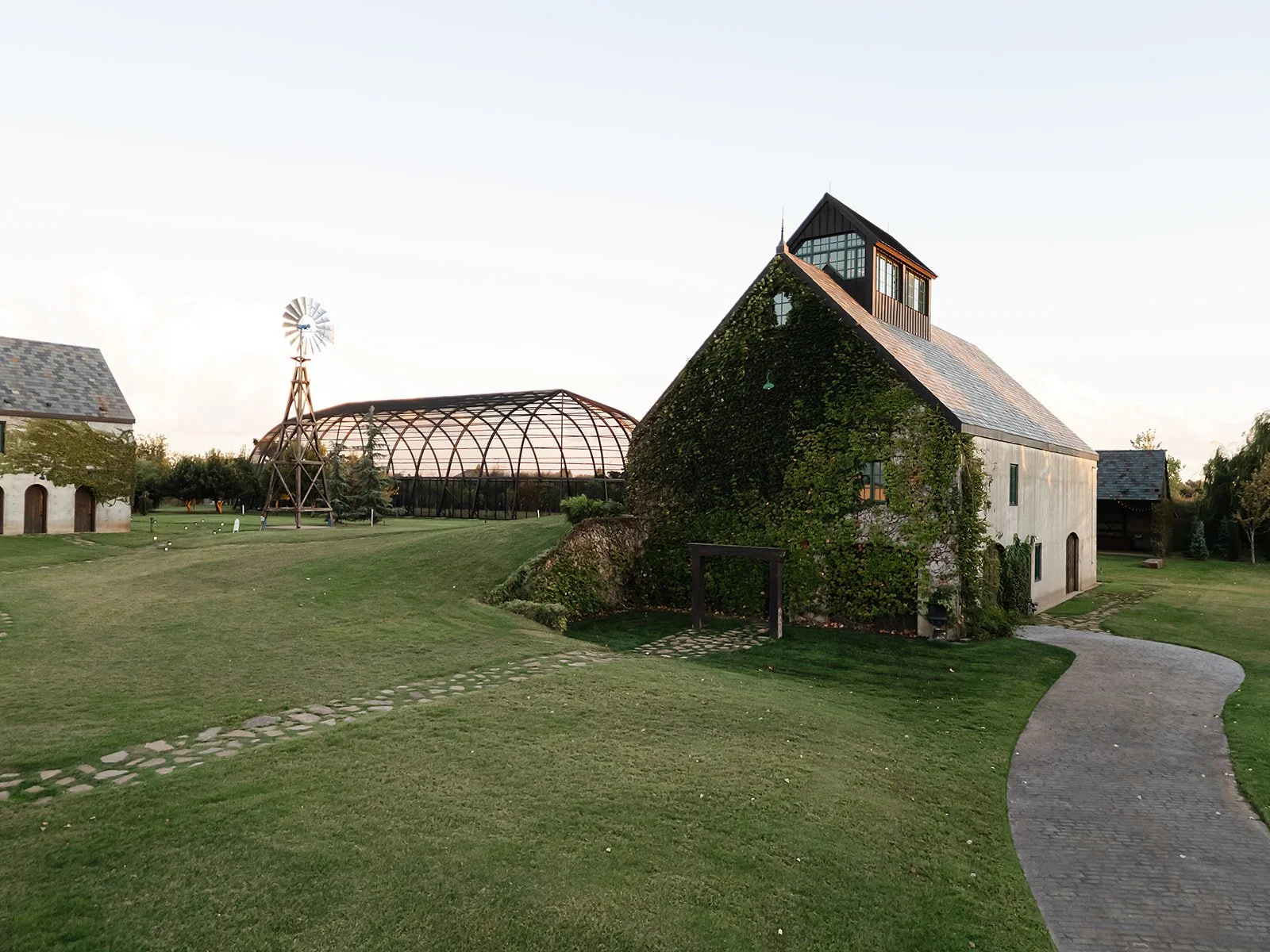 A rustic countryside scene with a vine-covered stone house, a curved stone pathway, a windmill, and a metal greenhouse structure in the background, under a clear sky.