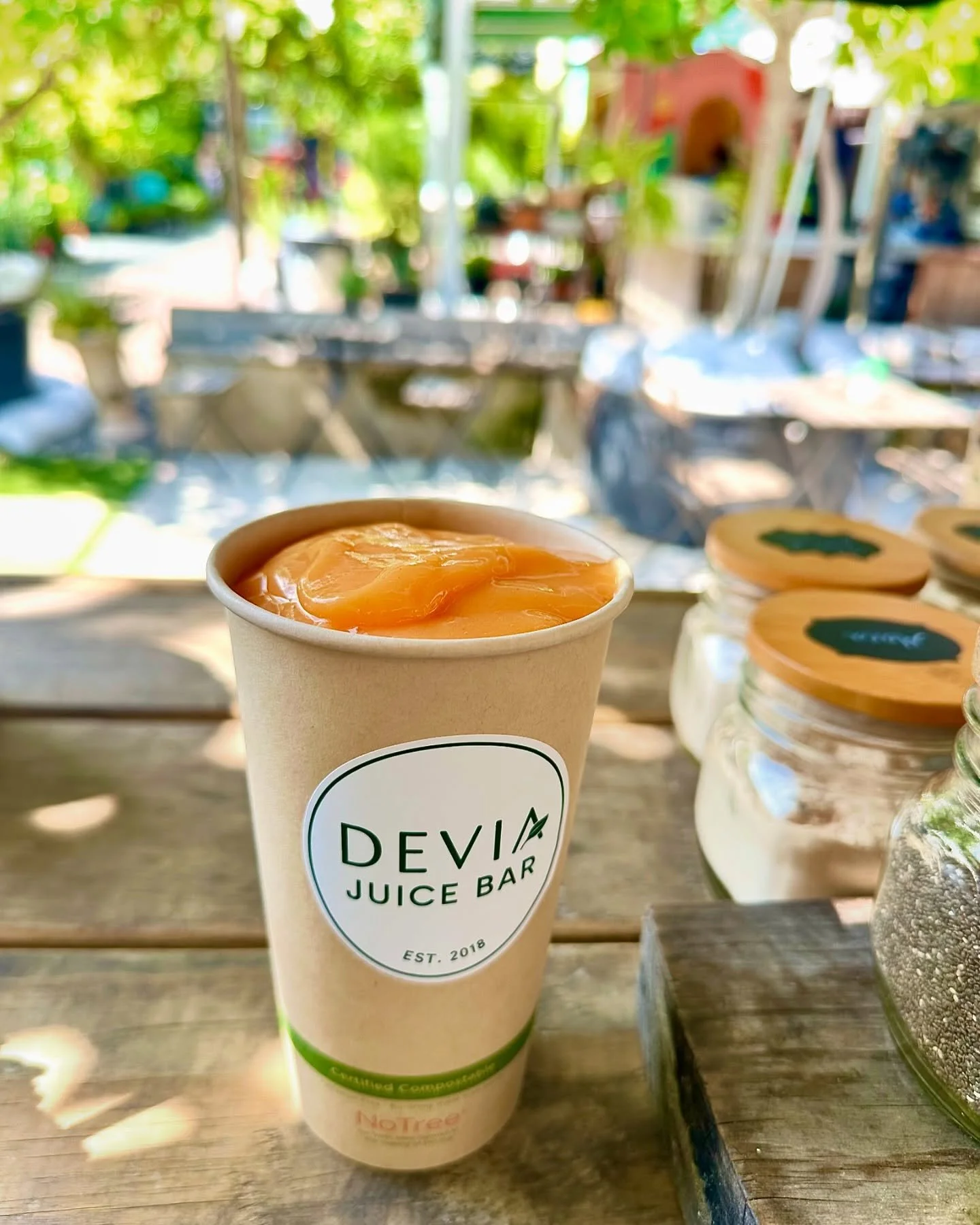 A paper cup labeled 'Devi Juice Bar' with fresh orange juice or smoothie on a wooden table outdoors, with jars of seeds and a blurred background of trees and outdoor seating.