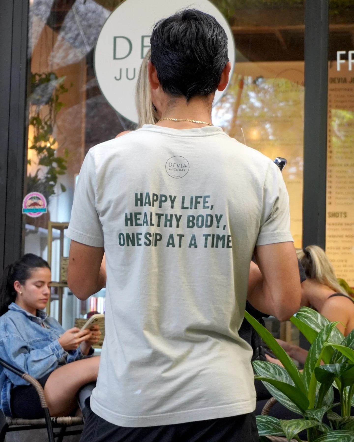 Person standing in front of a restaurant window with a t-shirt that reads, 'HAPPY LIFE, HEALTHY BODY, ONE SIP AT A TIME.' Inside, people are sitting and using their phones.