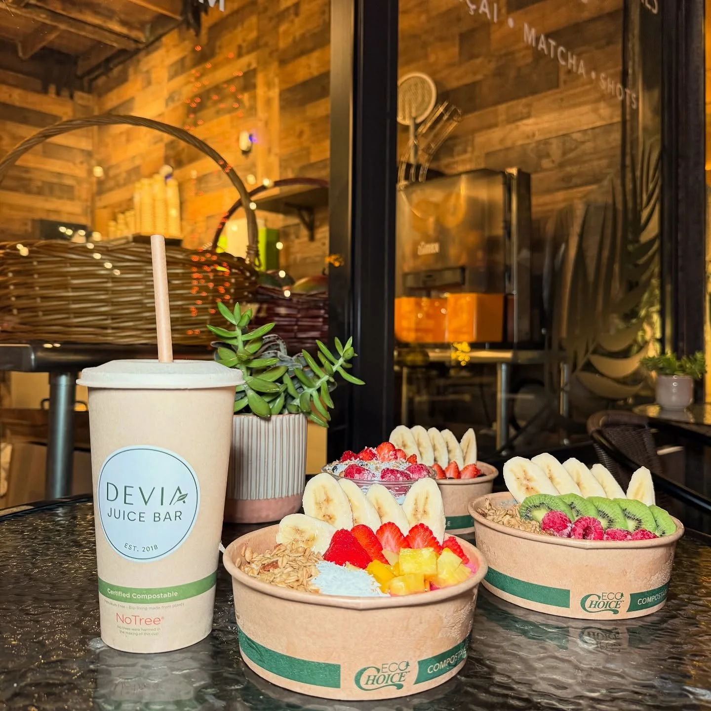 Three bowls of fresh fruit topped with sliced bananas, strawberries, raspberries, and other fruits on a glass table outside a juice bar, with a beige juice cup and potted succulent plant in the foreground.