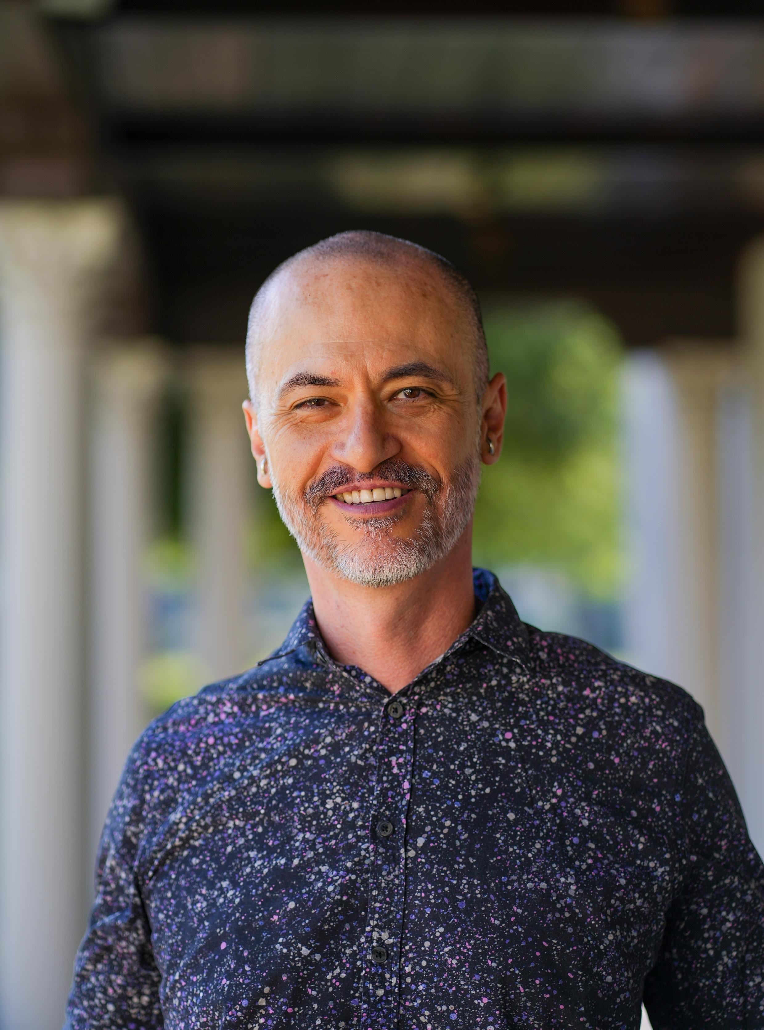 Close-up portrait of a smiling man with a bald head, gray beard, and earrings, wearing a dark shirt with a colorful speckled pattern, standing outdoors with blurred green background.