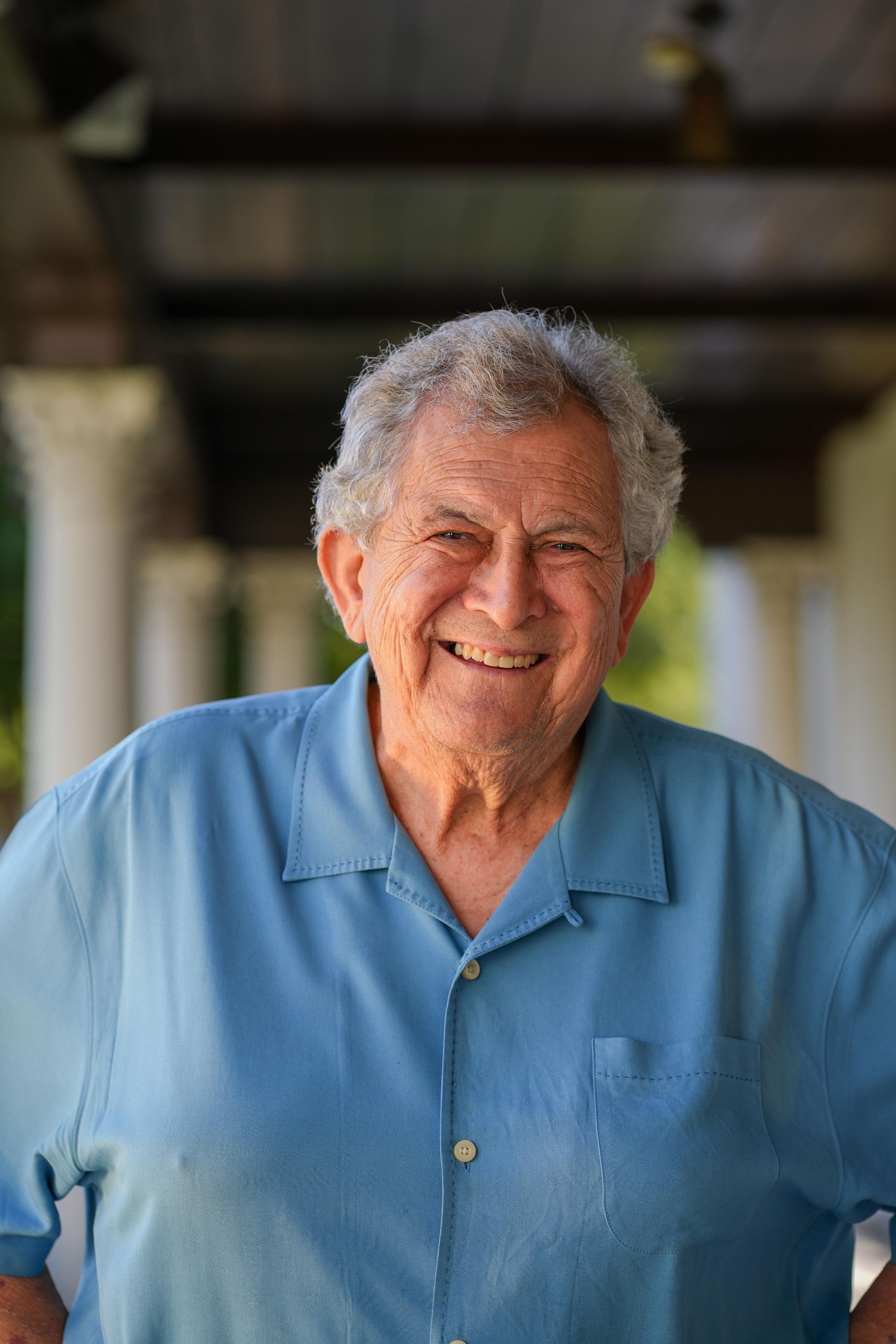Close-up of a smiling elderly man with gray hair, wearing a light blue button-up shirt, standing outdoors with a blurred background of trees and structure.