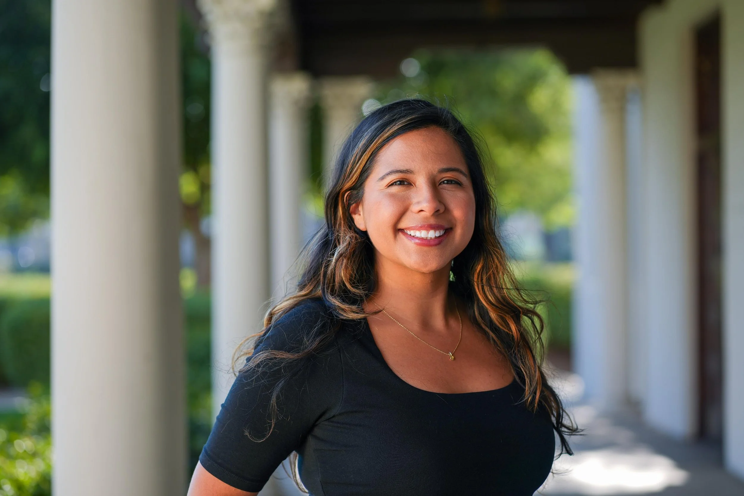 A woman with dark hair and highlights, smiling, wearing a black top, standing outdoors under a structure with white columns, with greenery and trees in the background.