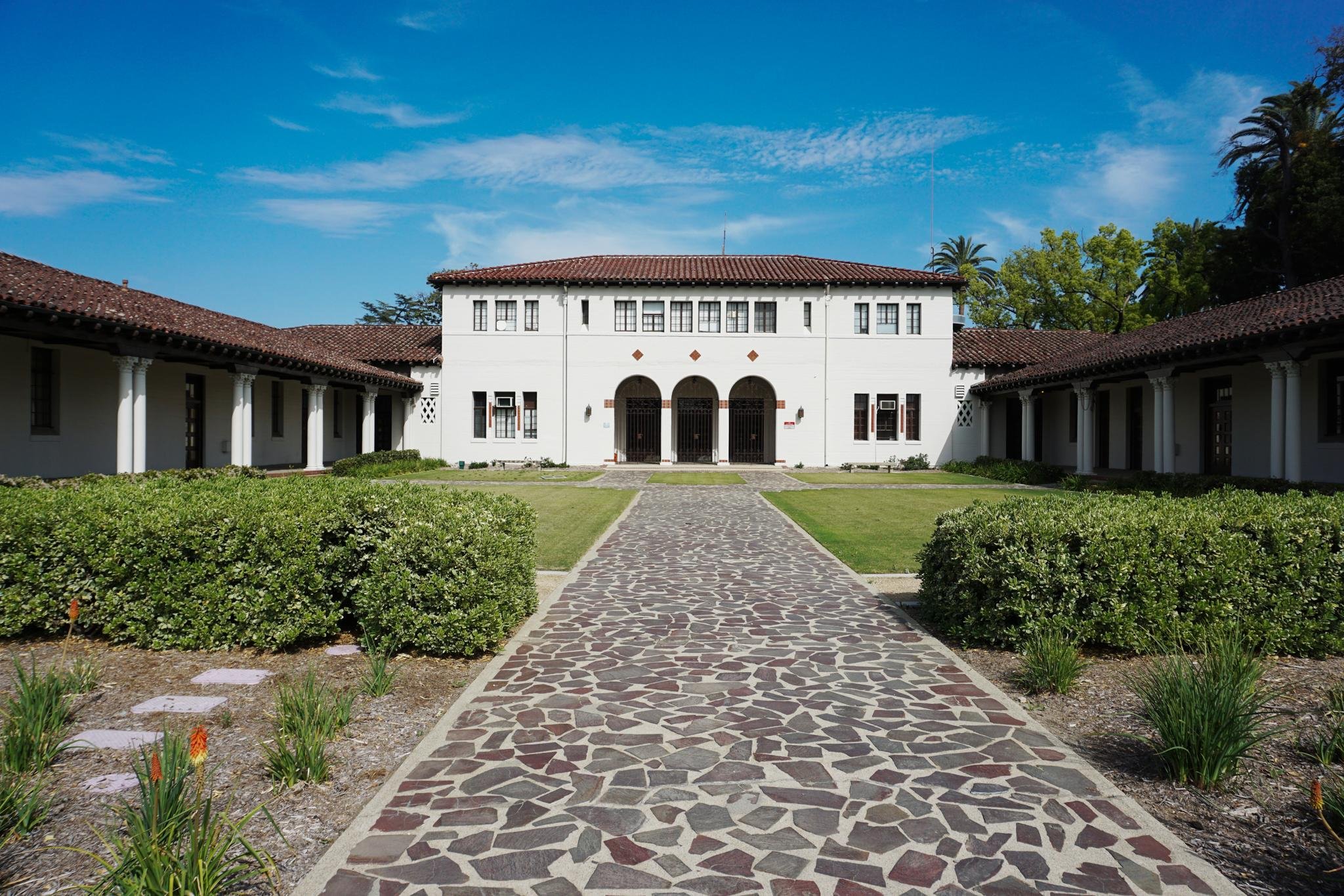 A large white building with a tiled red roof, arched entryways, and multiple windows, surrounded by greenery, with a stone pathway leading to the entrance under a blue sky.