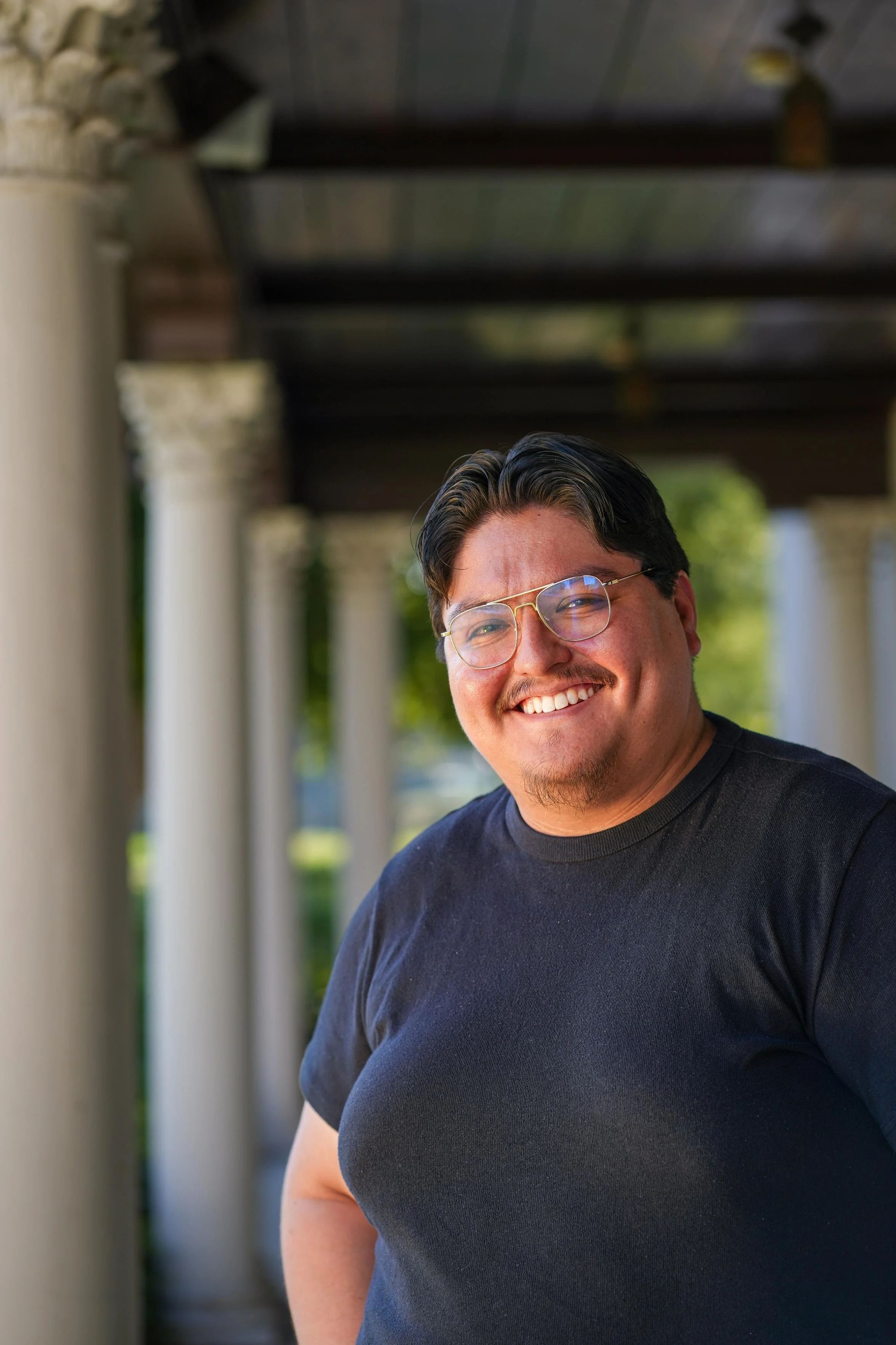 A smiling man wearing glasses and a black t-shirt, standing outdoors with a background of columns and greenery.