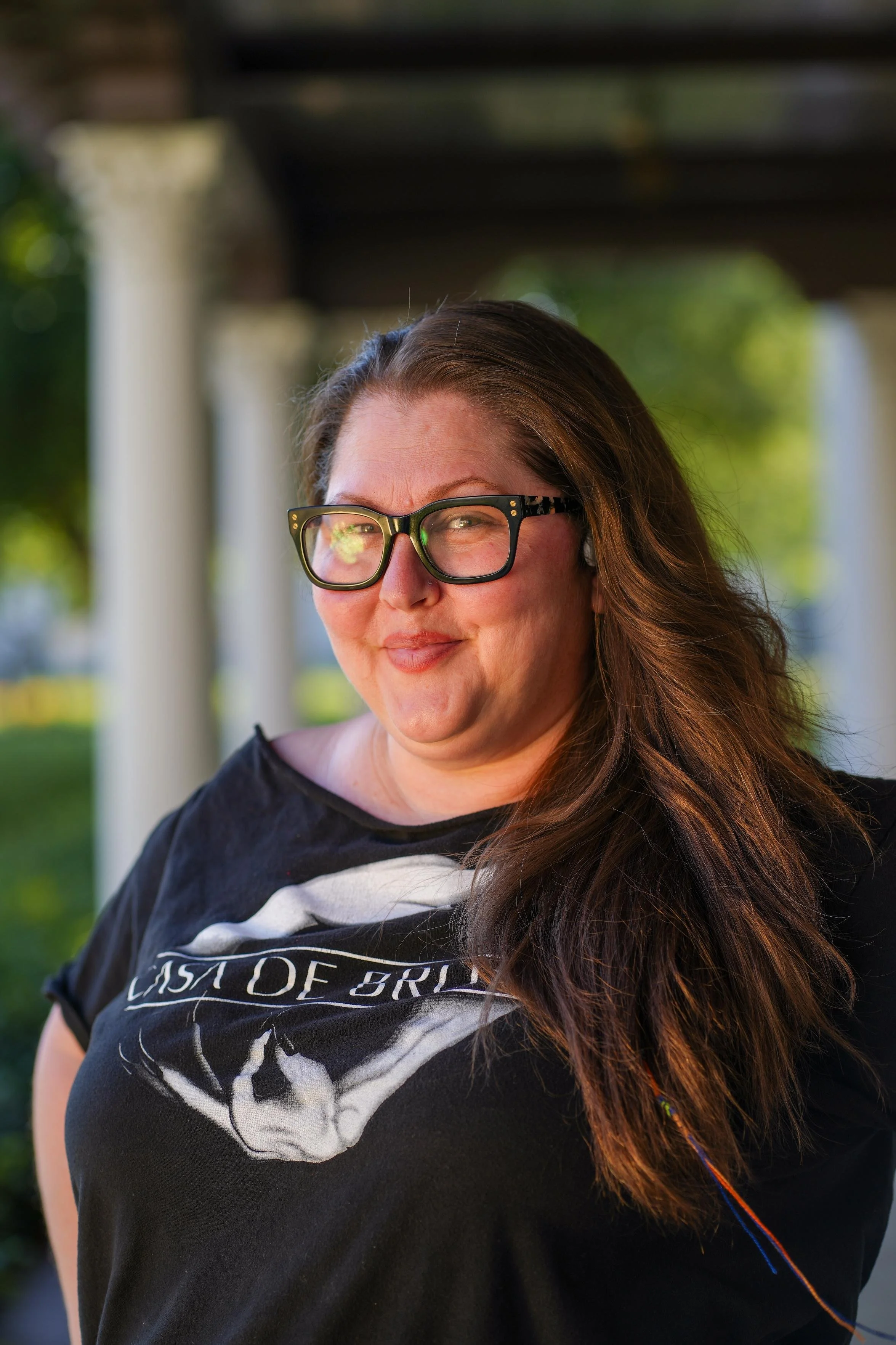 A woman with long brown hair, wearing large black glasses and a black t-shirt with a white graphic design, stands outdoors with blurred trees and columns in the background.