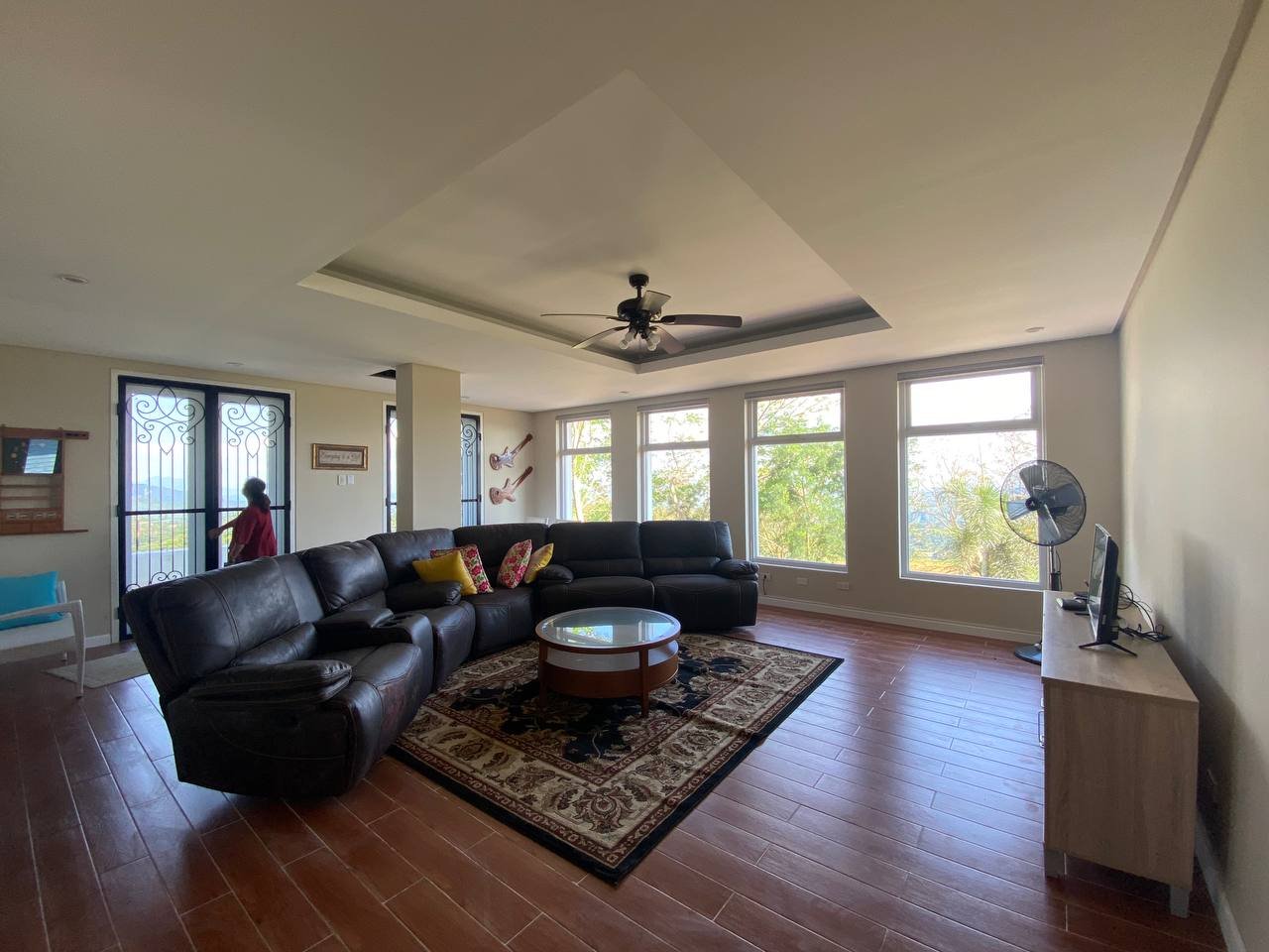 Living room with large sectional sofa, wooden coffee table, TV on a light-colored wood stand, ceiling fan, and large windows with views of trees outside.
