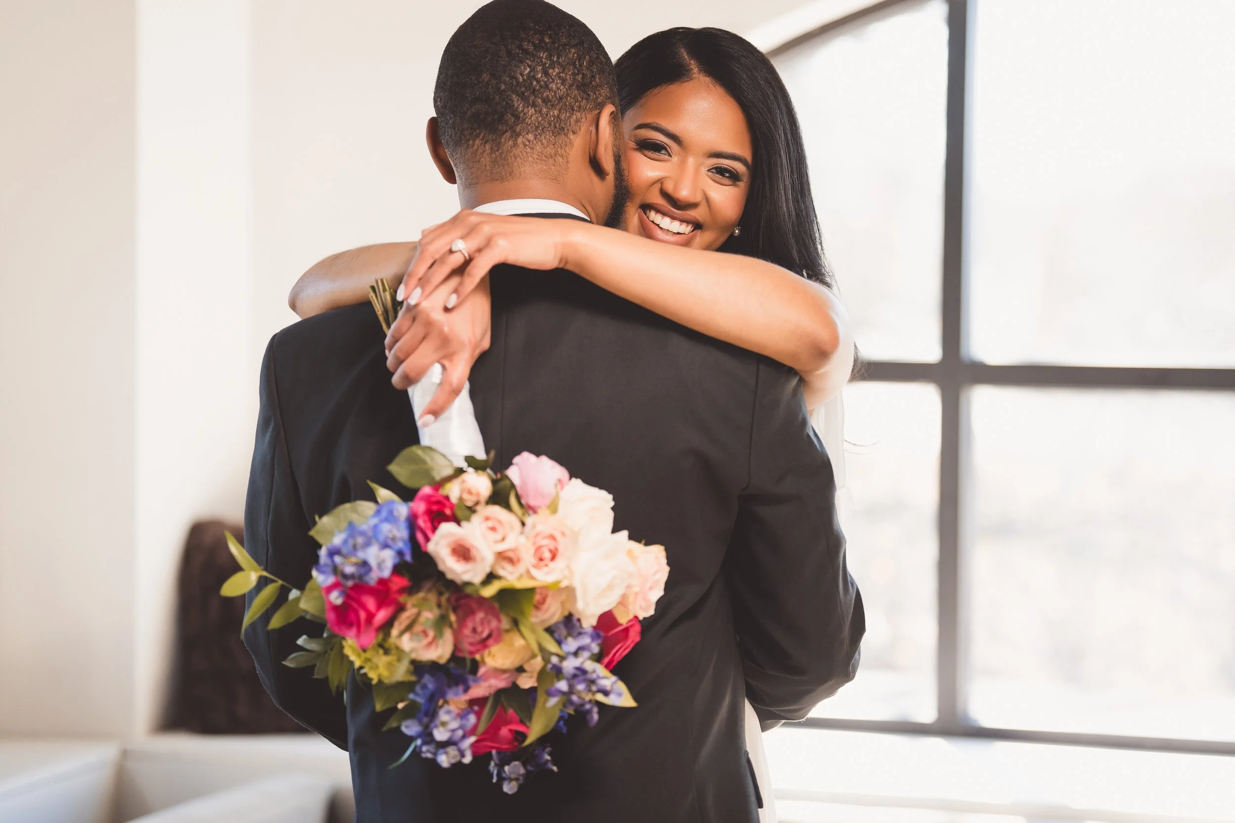 A woman hugging a man in a suit, holding a bouquet of colorful flowers, smiling indoors near a window.