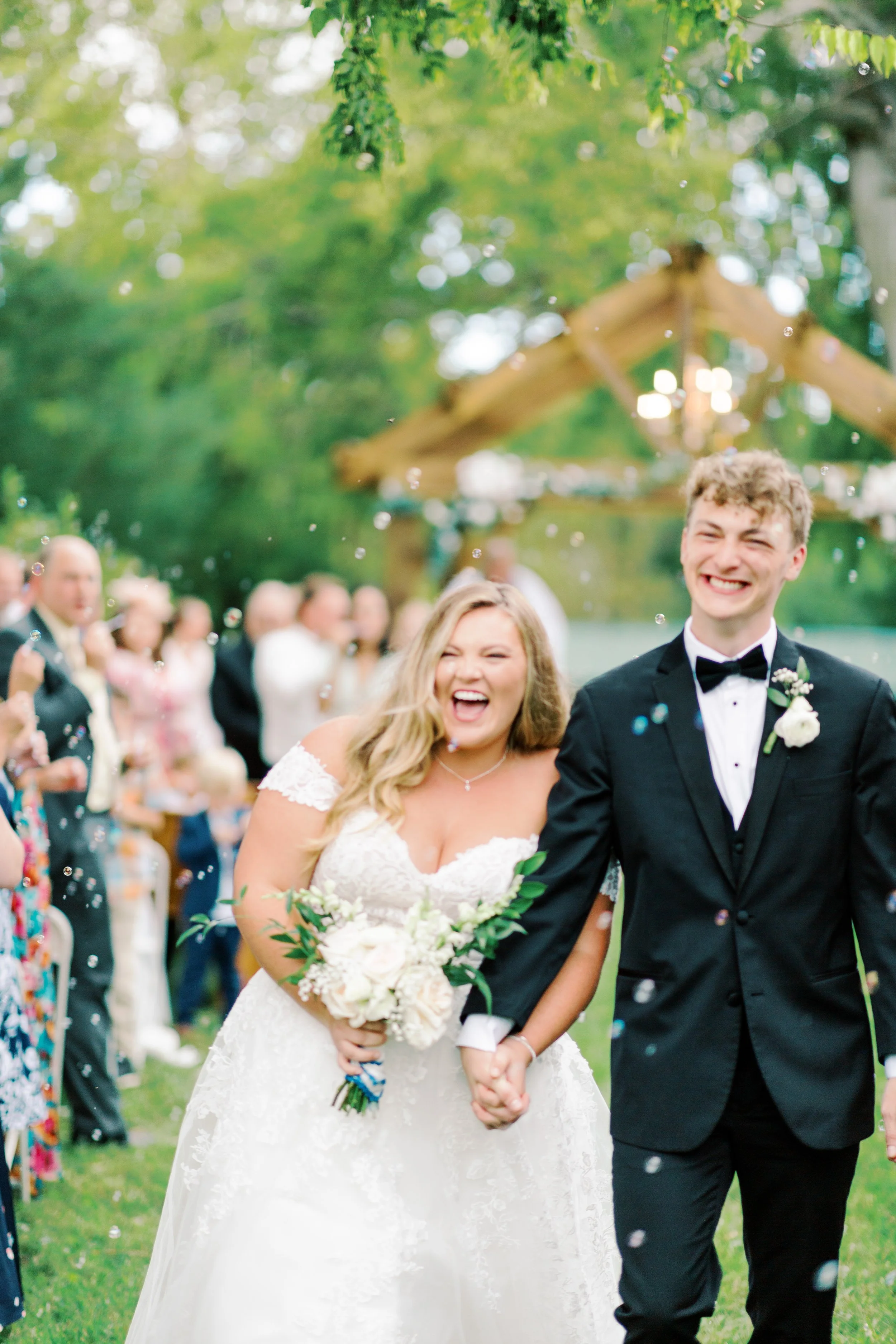 A bride and groom smiling and holding hands during an outdoor wedding, with guests celebrating in the background and confetti in the air.