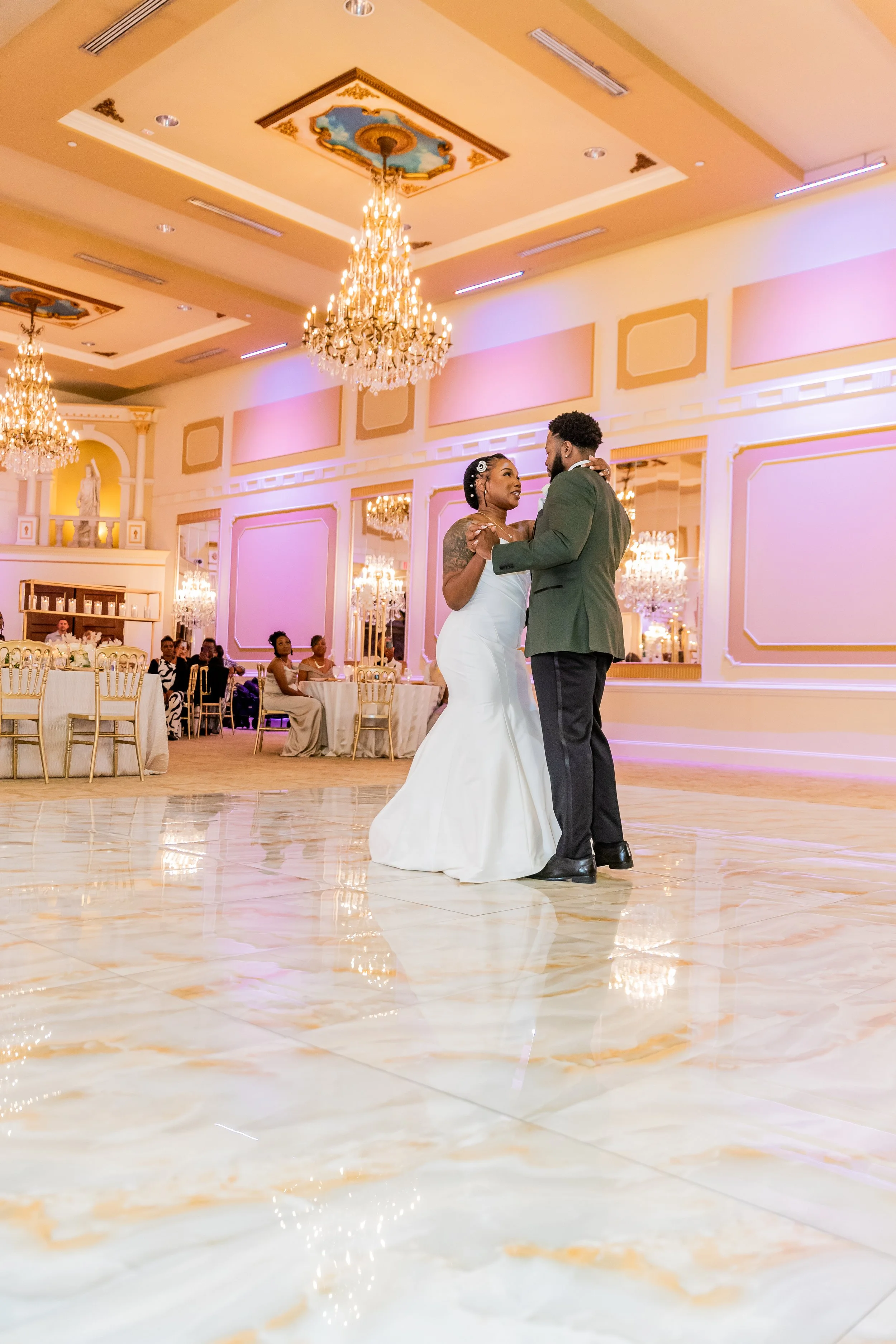 A bride and groom dance together at their wedding reception in a luxurious, decorated ballroom with chandeliers and other guests watching in the background.